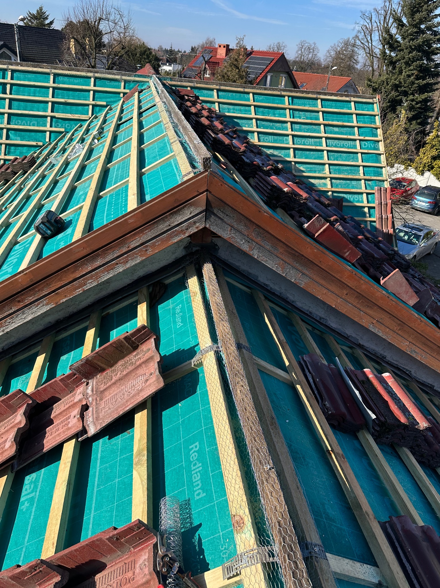 Fragment dachu w trakcie prac. Widoczna drewniana konstrukcja, membrana w kolorze turkusowym i częściowo ułożona dachówka w odcieniach czerwieni i brązu. Prace dekarskie w toku.