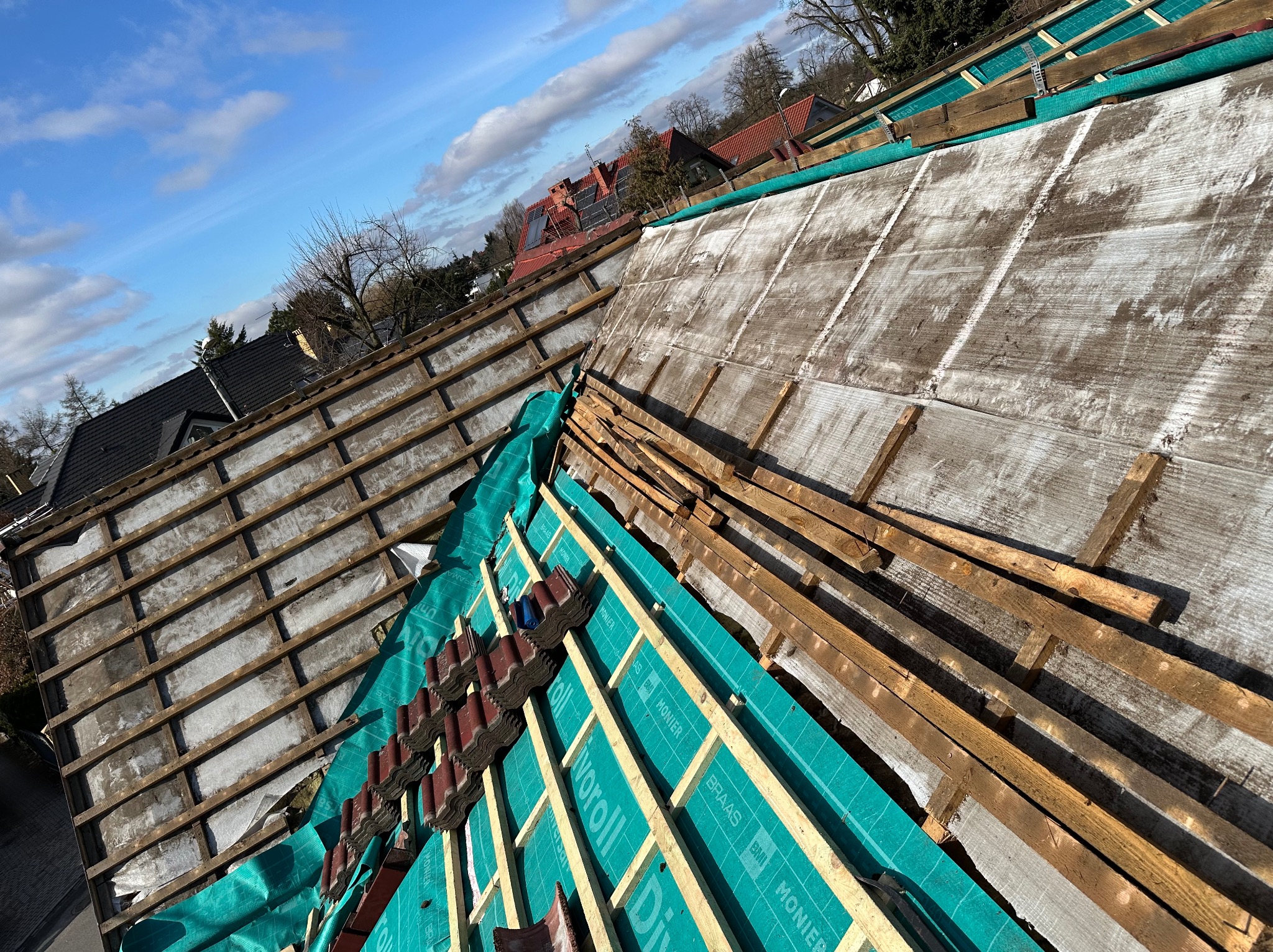 Dach w trakcie prac: widoczna konstrukcja, membrana w kolorze turkusowym z nadrukiem, częściowo ułożona dachówka. Słoneczny dzień, błękitne niebo z chmurami.