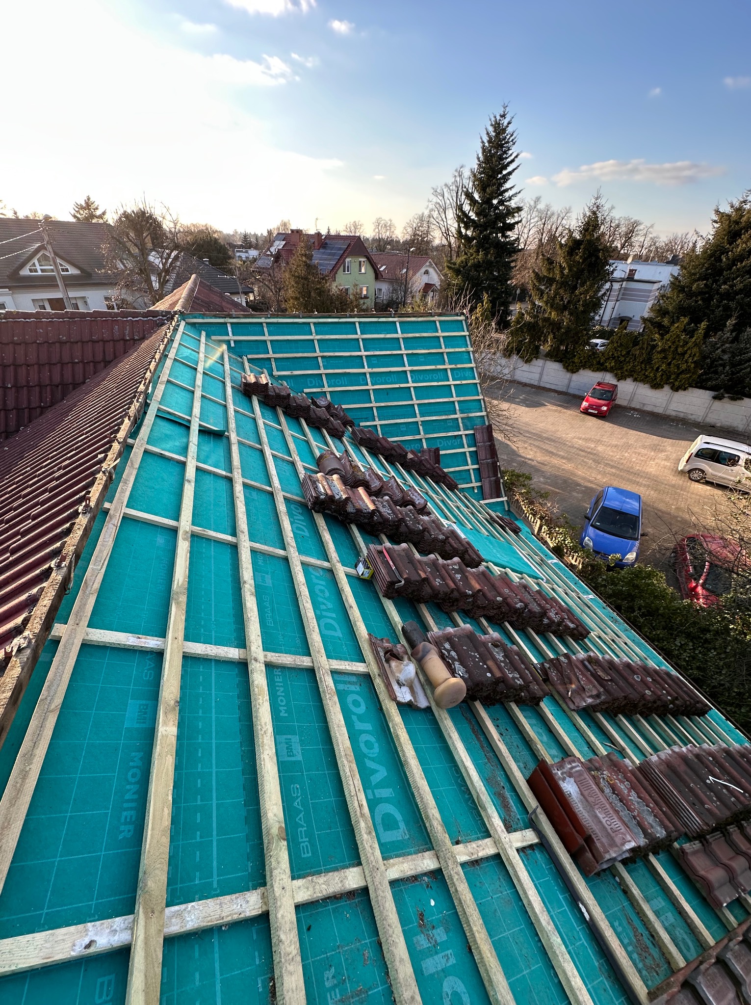 Układanie dachówki na łatach na folii paroizolacyjnej z widocznymi napisami producenta. Częściowo pokryty dach z perspektywy z góry, w tle zabudowa jednorodzinna.