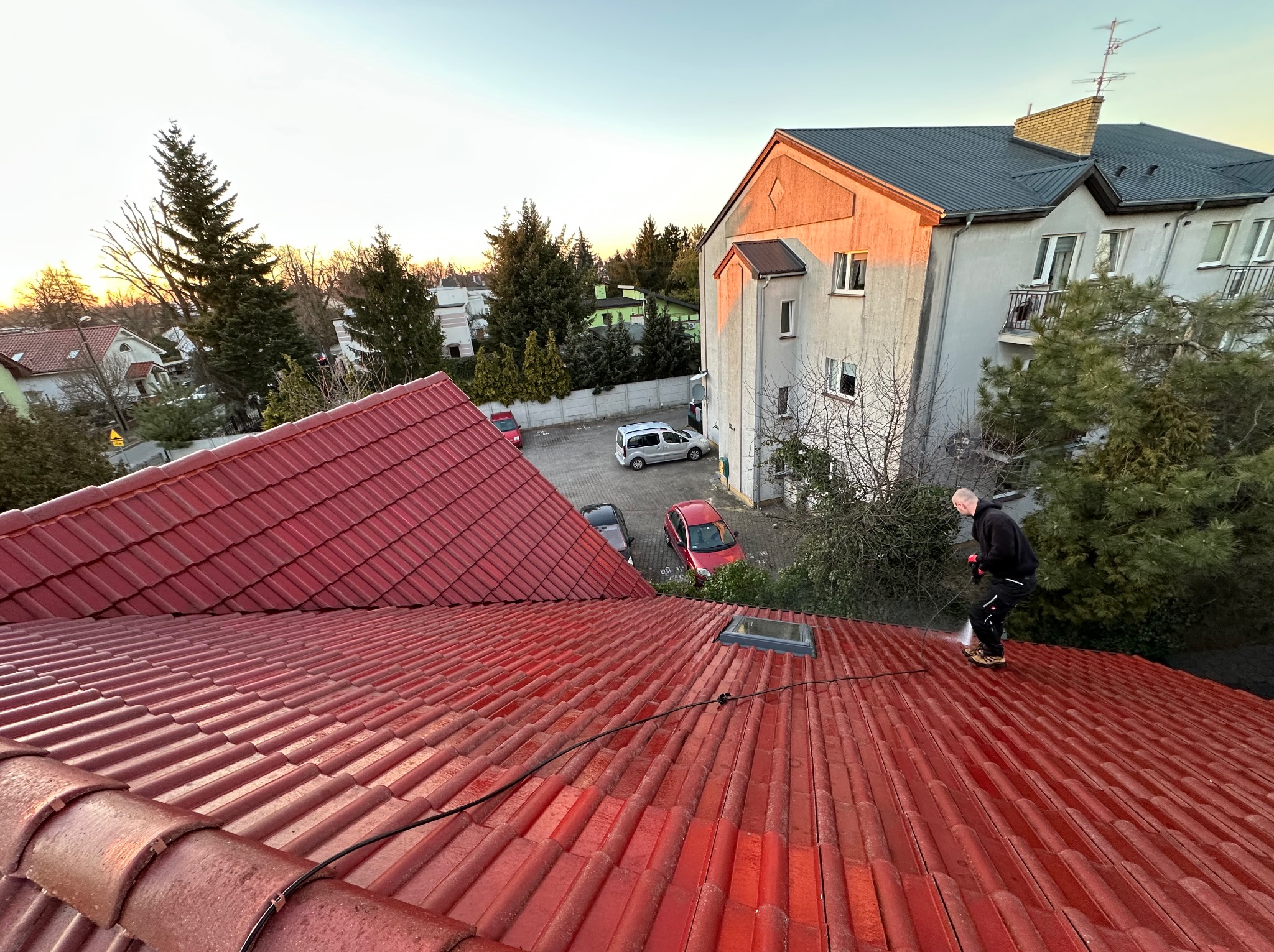 Czerwony dach z dachówki ceramicznej czyszczony myjką ciśnieniową przez dekarza. W tle budynek mieszkalny i drzewa w promieniach zachodzącego słońca.