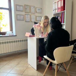 Nowa Dieta Katarzyna Nowak - Konsultacja: blondynka w okularach omawia plan z klientką przy białym biurku w jasnym gabinecie z dyplomami i regałem na dokumenty w tle.