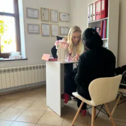 Konsultacja: blondynka w okularach omawia plan z klientką przy białym biurku w jasnym gabinecie z dyplomami i regałem na dokumenty w tle.