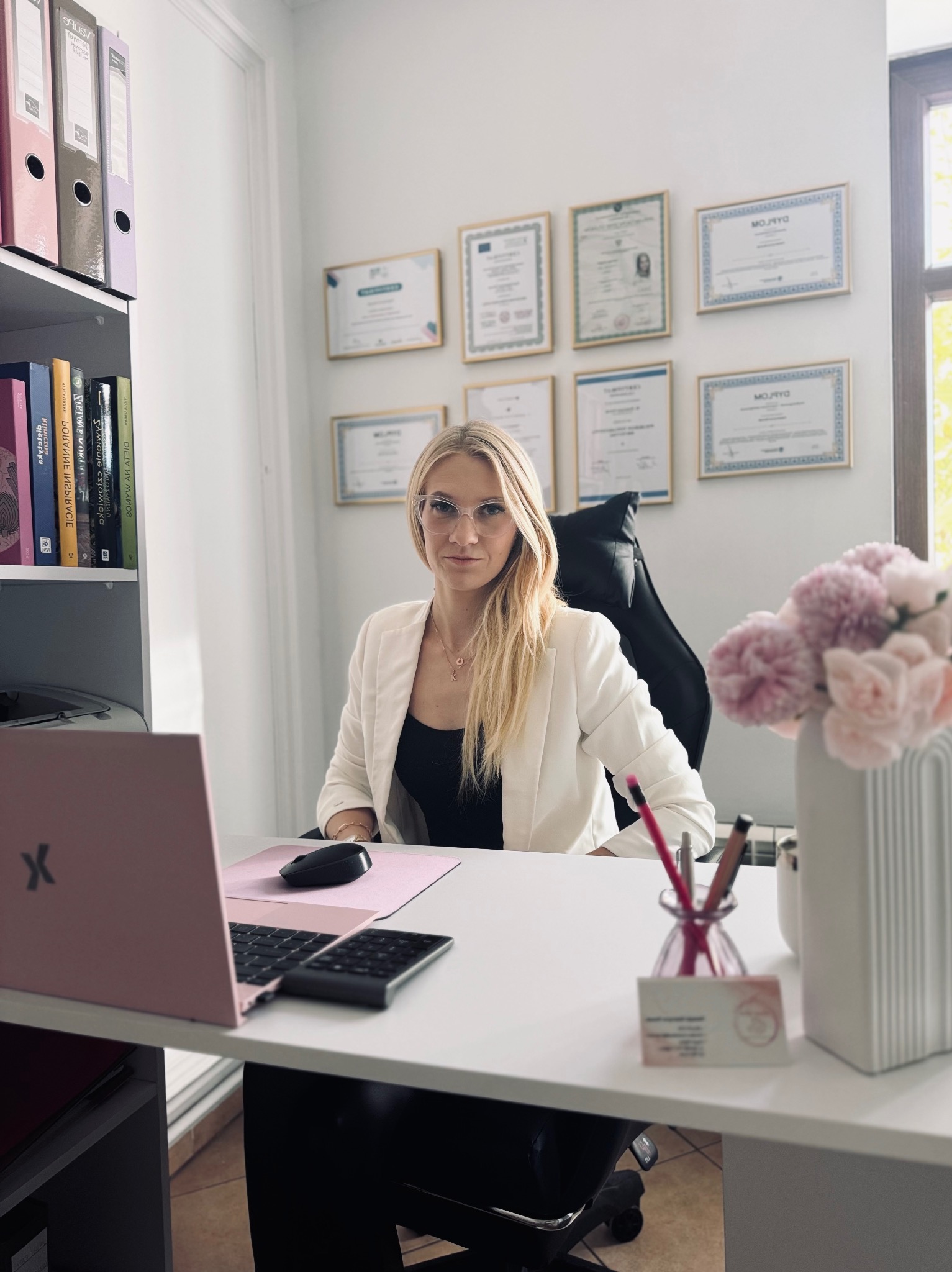 Profesjonalny gabinet: blondynka w okularach siedzi za biurkiem z laptopem, kwiatami i certyfikatami na ścianie. Jasne, naturalne światło.