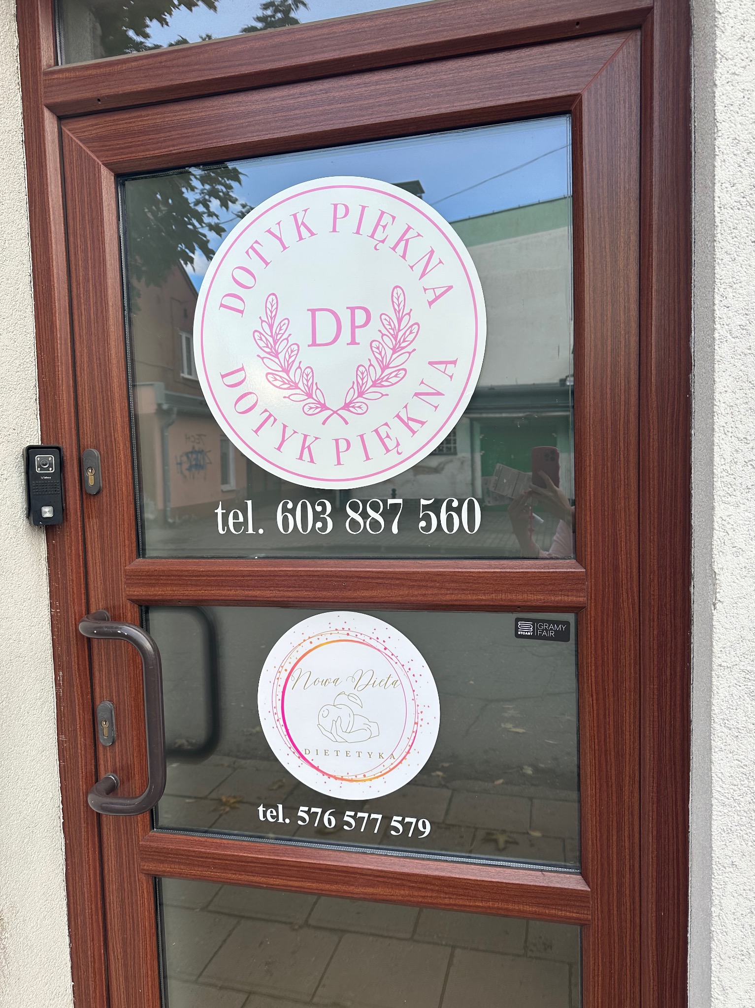 Brązowe drzwi wejściowe z naklejonymi okrągłymi reklamami: „Dotyk Piękna” i „Nowa Dieta Dietetyk” z numerami telefonów. Widoczny fragment chodnika i budynków w tle.