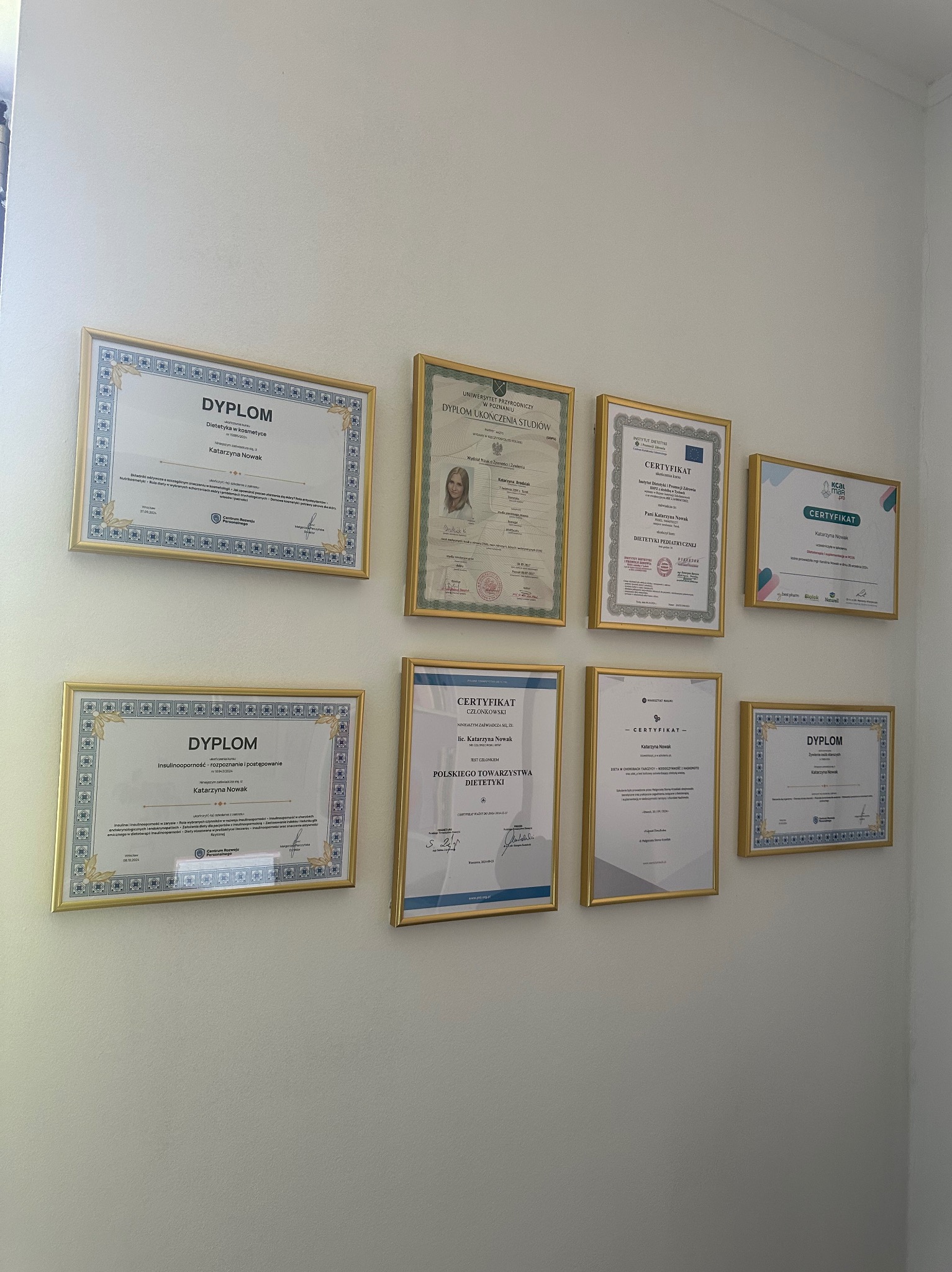 Ściana z dyplomami i certyfikatami w złotych ramkach, potwierdzającymi kwalifikacje zawodowe, w tym dyplom z dietetyki i kosmetyki Katarzyny Nowak.