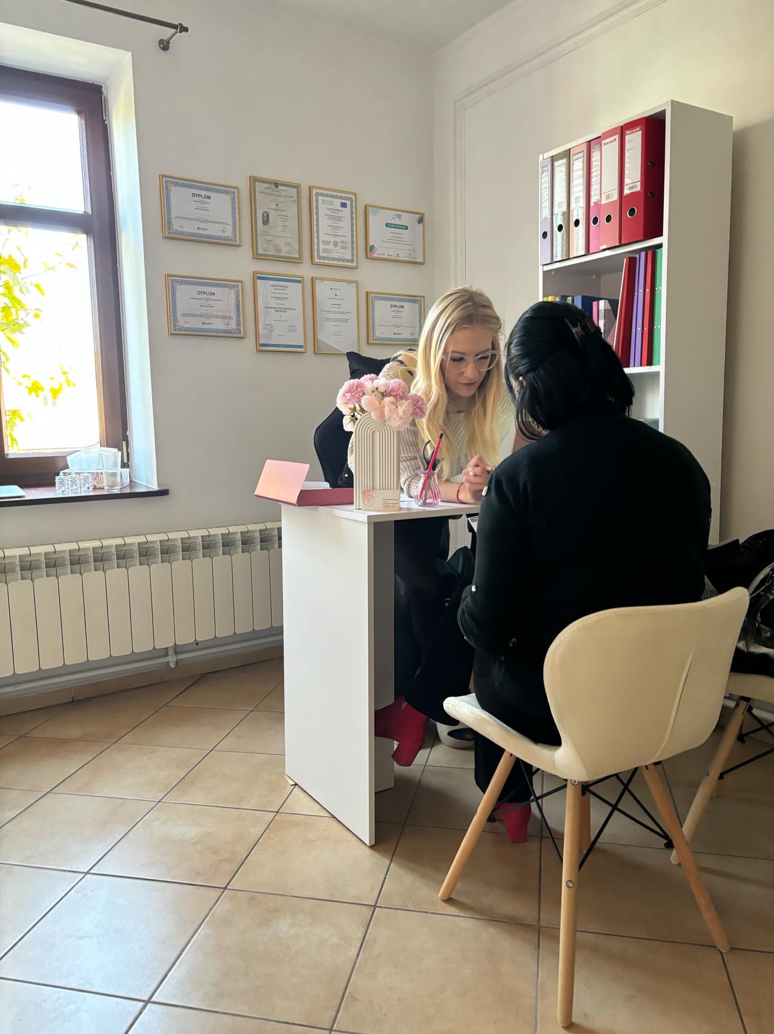 Konsultacja: blondynka w okularach omawia plan z klientką przy białym biurku w jasnym gabinecie z dyplomami i regałem na dokumenty w tle.