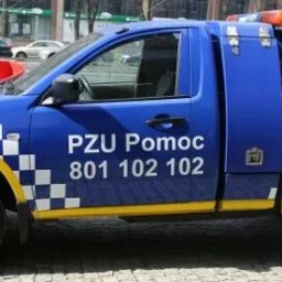 MIECZYSŁAW FICNER - Niebiesko-żółty samochód pomocy drogowej PZU Pomoc z numerem telefonu 801 102 102 na tle miejskiego krajobrazu. Pojazd z dźwigiem holowniczym.