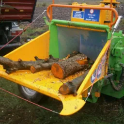 Zielono-żółty rębak Heizomat HM 6-300 z załadowanymi gałęziami i pniem drzewa, widoczny fragment ciężarówki w tle.