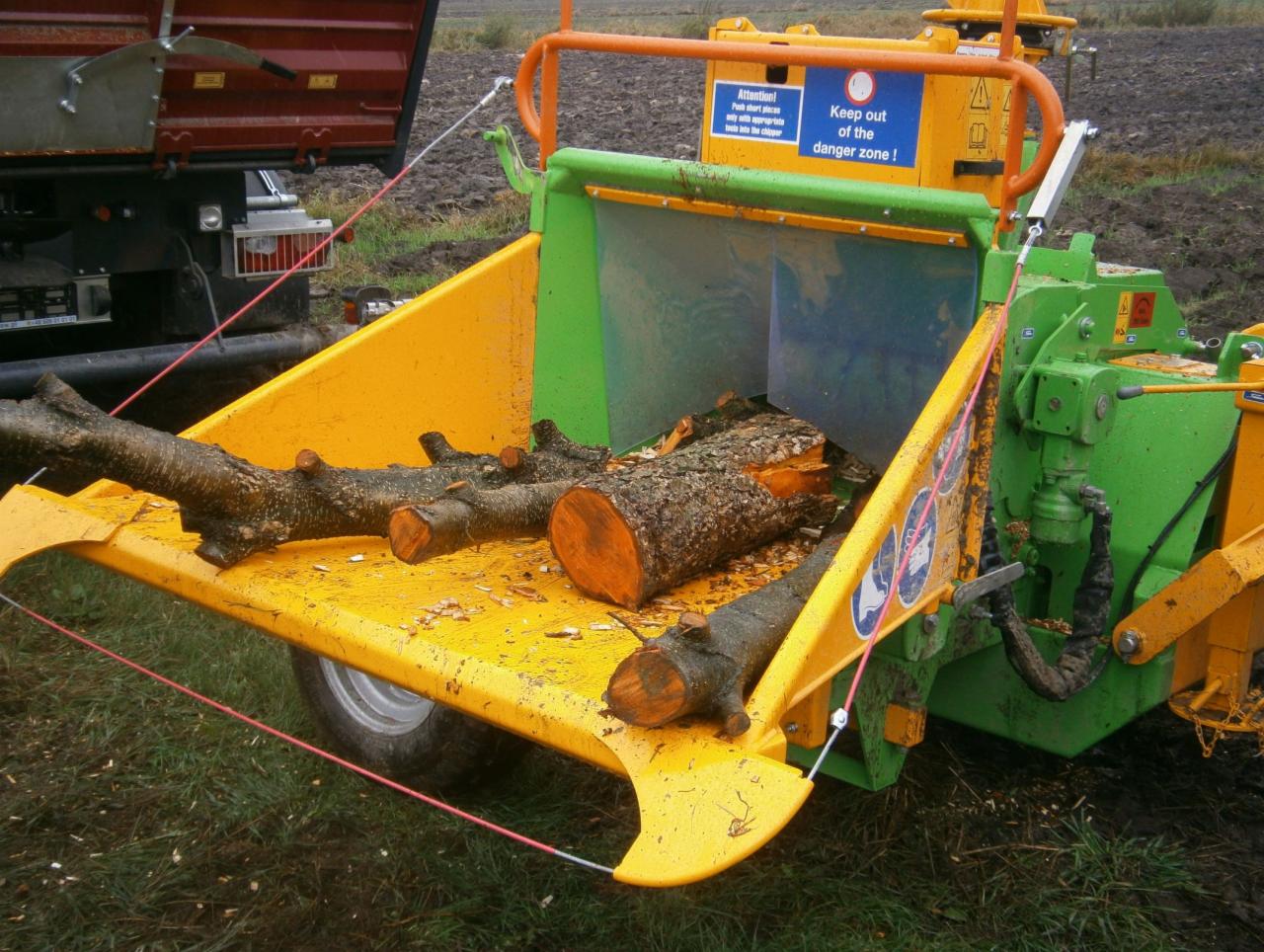 Zielono-żółty rębak Heizomat HM 6-300 z załadowanymi gałęziami i pniem drzewa, widoczny fragment ciężarówki w tle.