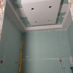 Firma Ogólnobudowlane Krzysztof Pawłowski - Sufit podwieszany z płyt gipsowo-kartonowych z otworami na oświetlenie, częściowo wyszpachlowany, z żółtą poziomicą opartą o ścianę. W tle surowy mur.