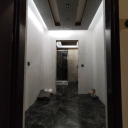 Firma Ogólnobudowlane Krzysztof Pawłowski - Wykończony korytarz z czarną marmurową podłogą, szarymi ścianami i podwieszanym sufitem z oświetleniem LED. Widok zza futryny drzwi.