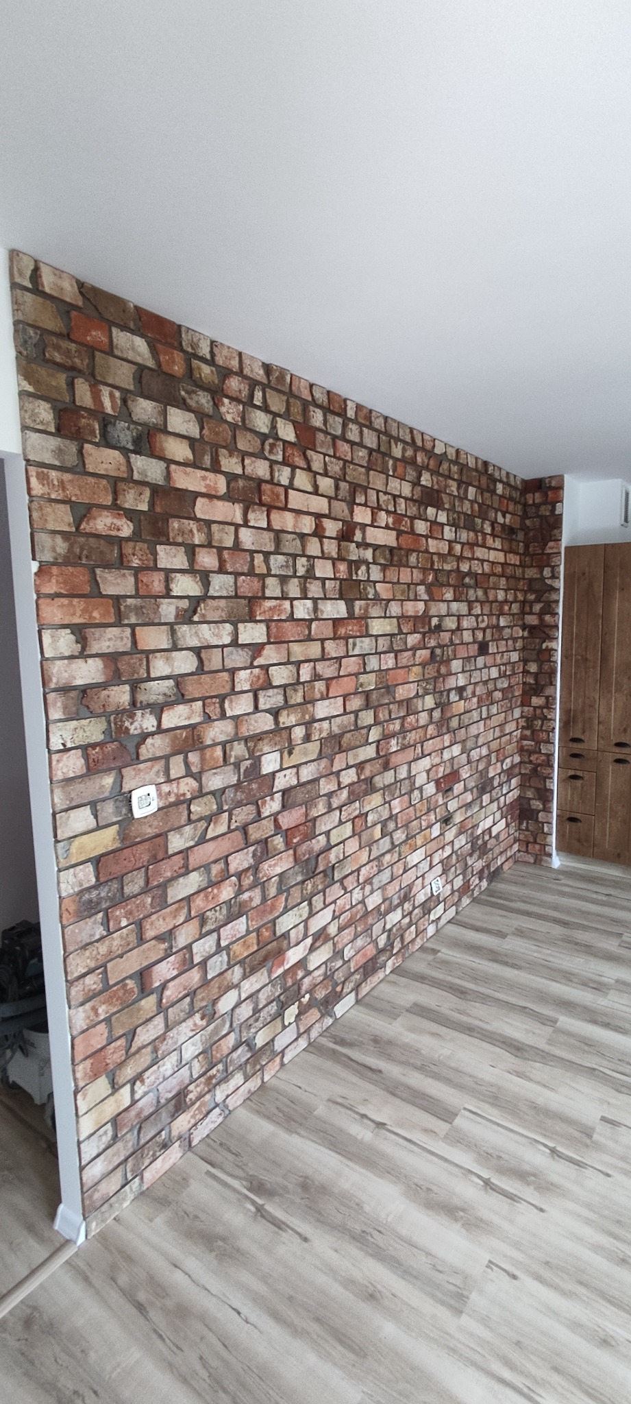 Ściana wykończona cegłą dekoracyjną w odcieniach brązu i beżu, z widocznym gniazdkiem elektrycznym. Podłoga z paneli imitujących drewno. Całość w jasnym pomieszczeniu.
