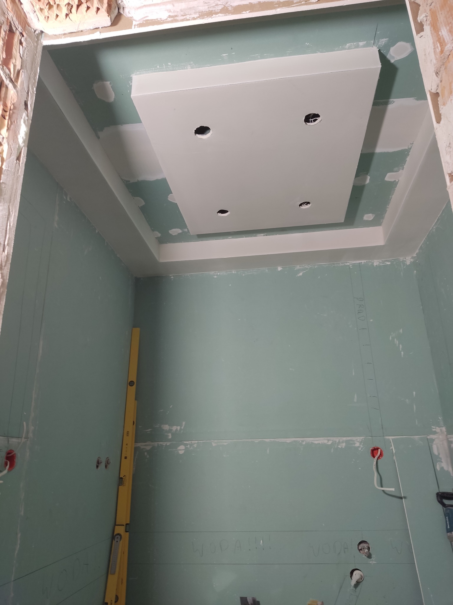 Sufit podwieszany z płyt gipsowo-kartonowych z otworami na oświetlenie, częściowo wyszpachlowany, z żółtą poziomicą opartą o ścianę. W tle surowy mur.