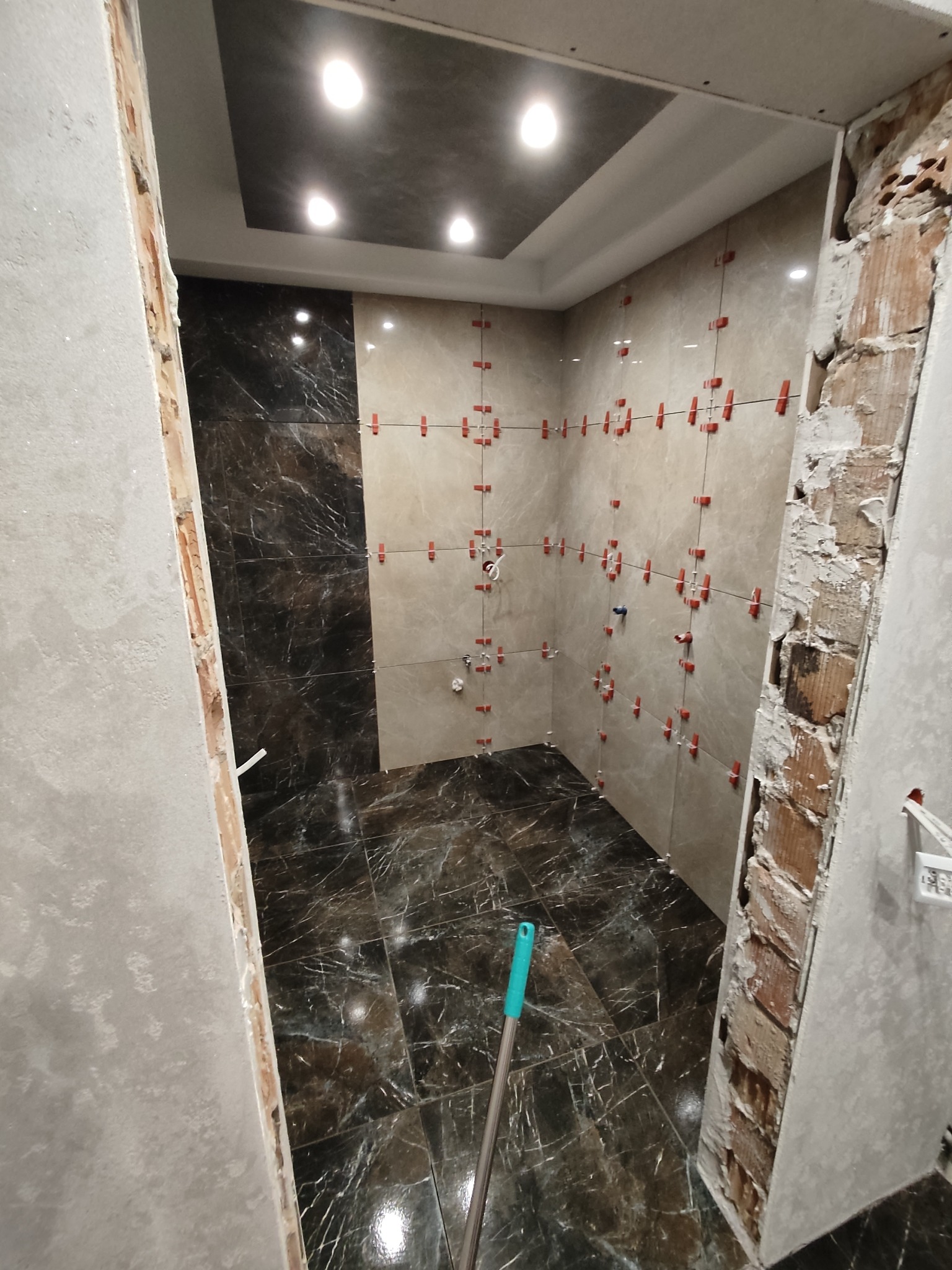Wnętrze łazienki w trakcie remontu: częściowo ułożone marmurowe płytki na podłodze i ścianach, widoczne klipsy poziomujące. Surowy stan ścian przy wejściu.