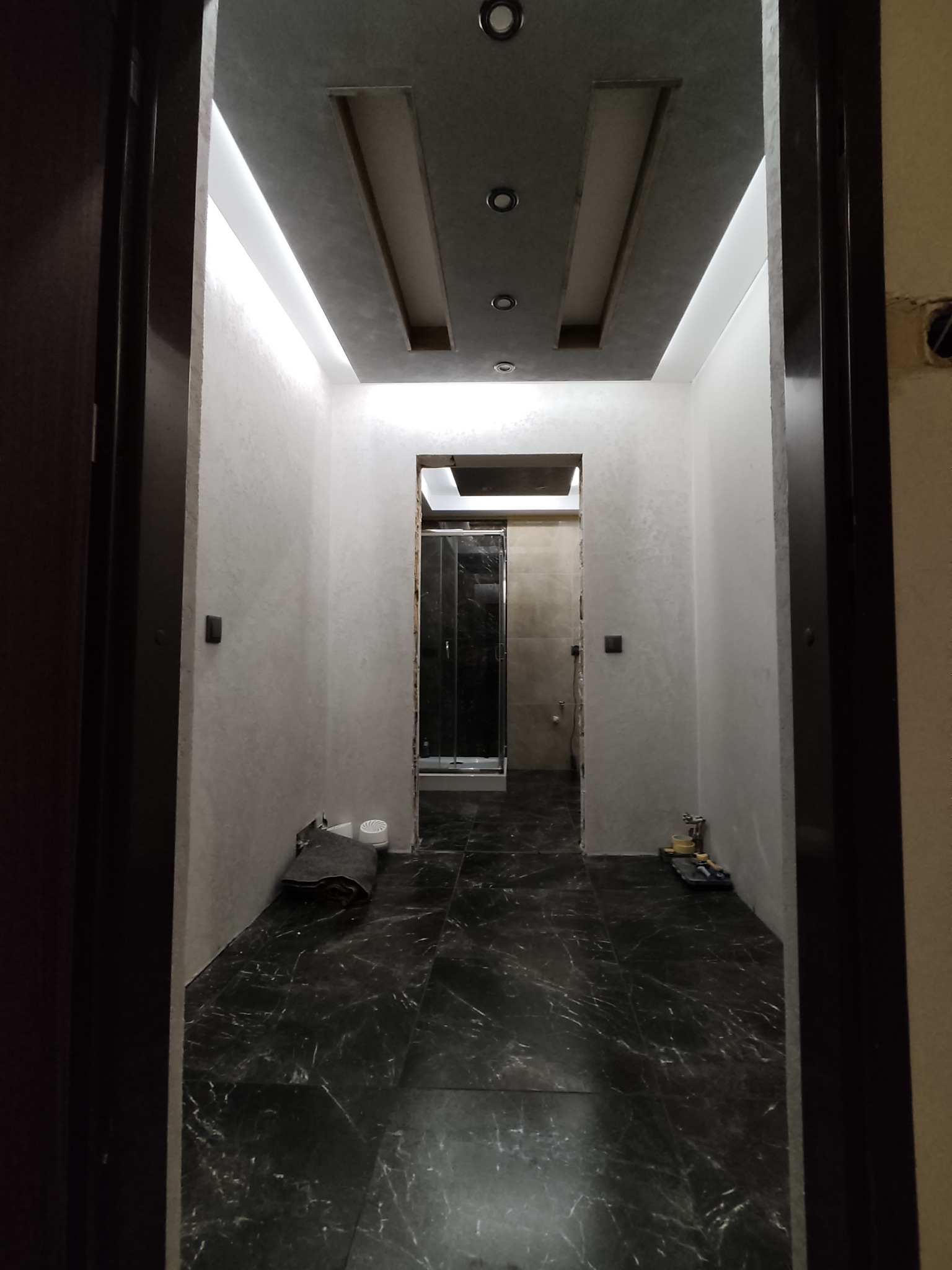 Wykończony korytarz z czarną marmurową podłogą, szarymi ścianami i podwieszanym sufitem z oświetleniem LED. Widok zza futryny drzwi.