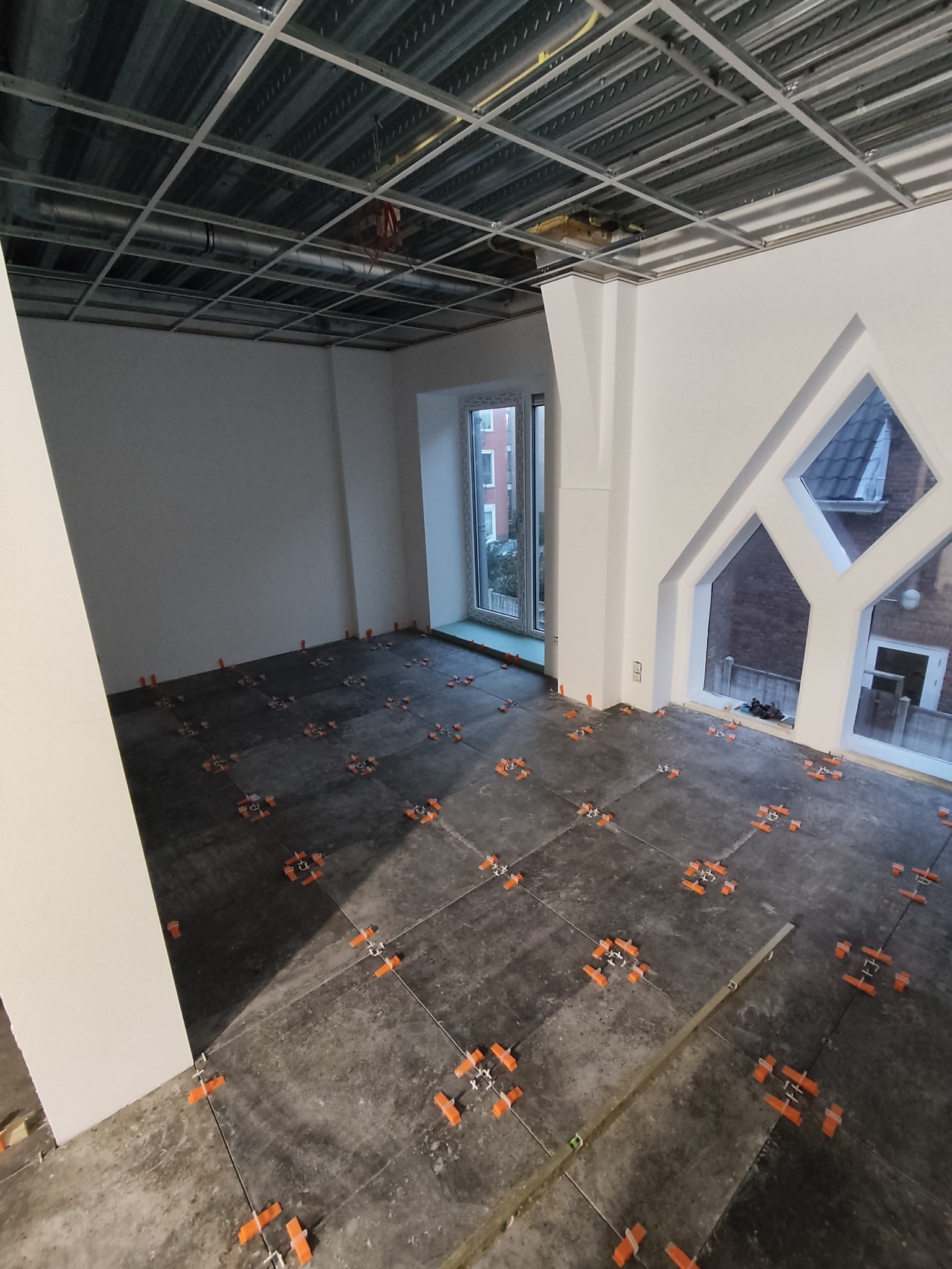 Układanie płytek podłogowych w jasnym wnętrzu z surowym stropem. Pomarańczowe krzyżyki dystansowe, okno z geometrycznym wykończeniem.