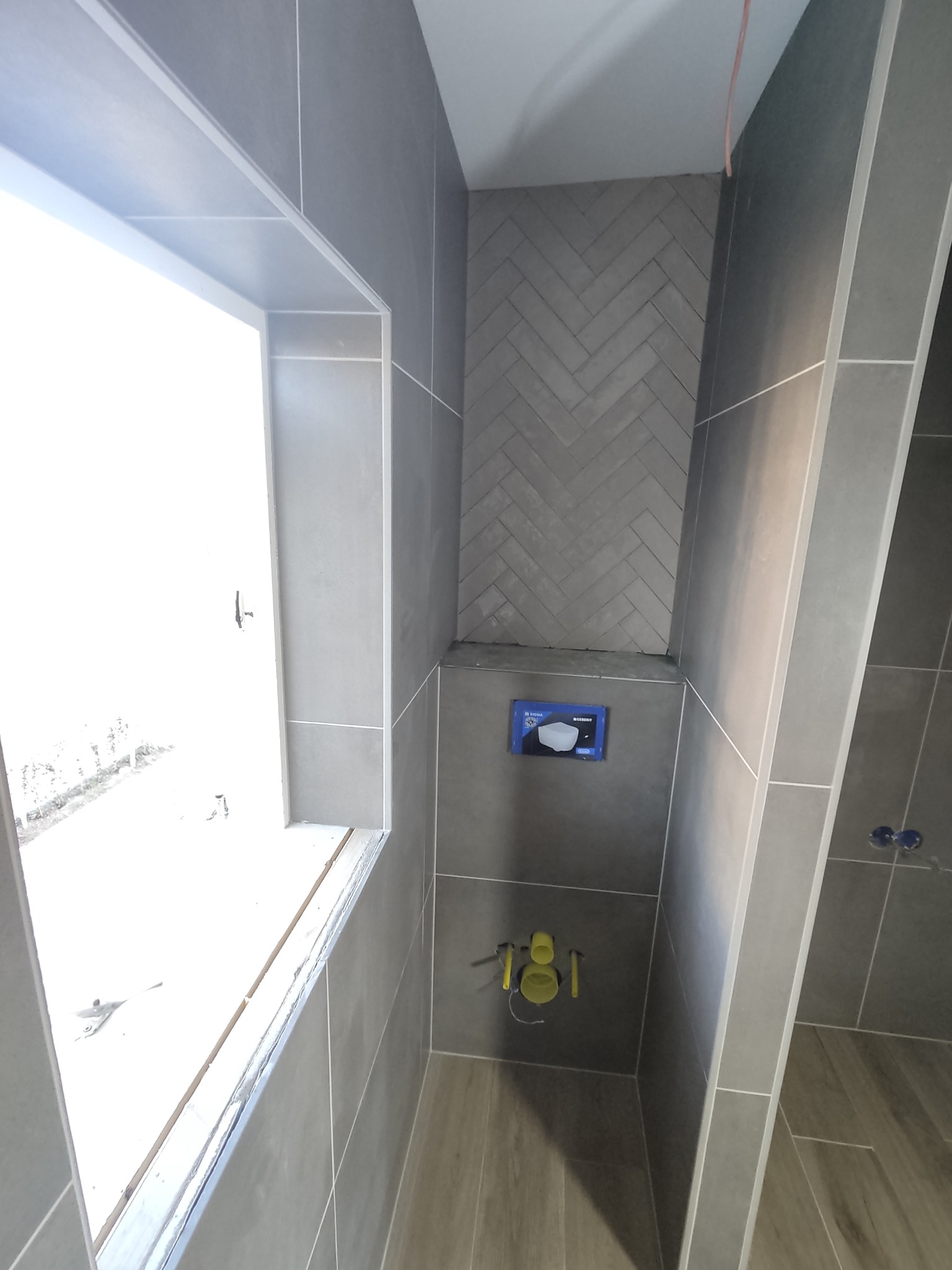 Szare płytki ułożone w jodełkę na ścianie łazienki, widoczne instalacje podtynkowe i okno z uszczelnieniem. Minimalistyczny design.