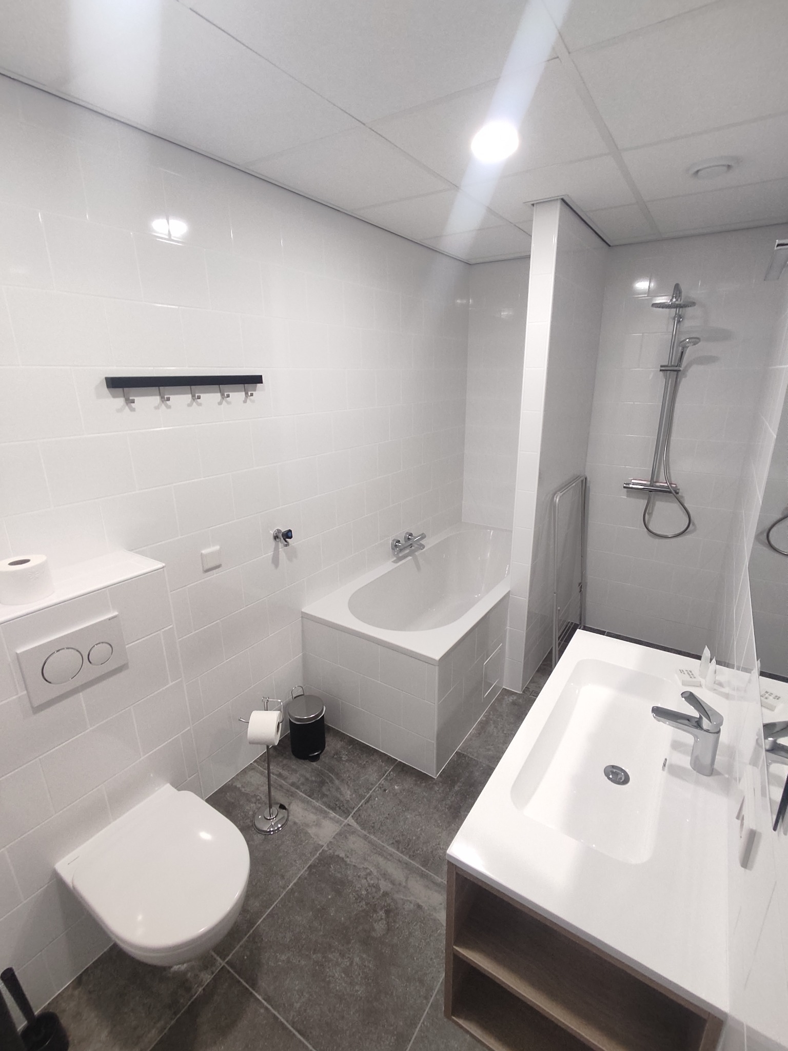 Nowoczesna, jasna łazienka z białymi płytkami, wiszącą toaletą, wanną i umywalką z szafką. Minimalistyczny design z czarnymi akcentami, np. wieszak na ręczniki.