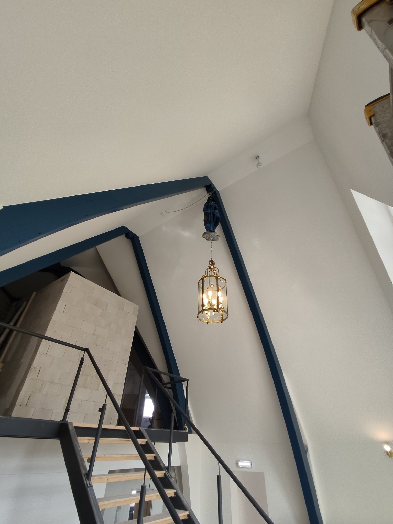 Wnętrze w trakcie wykańczania: białe ściany, niebieskie belki, schody z czarną balustradą i wisząca, ozdobna lampa. Widoczne materiały budowlane i drabina.