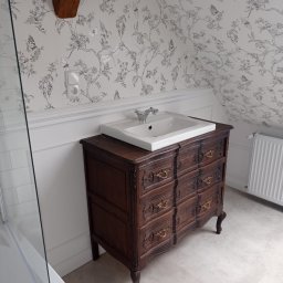 USŁUGI REMONTOWO-BUDOWLANE BIELAWSKI KRZYSZTOF MIROSŁAW - Umywalka nablatowa na odrestaurowanej, ciemnobrązowej komodzie z ozdobnymi uchwytami w łazience na poddaszu z białą tapetą w motywy roślinne.