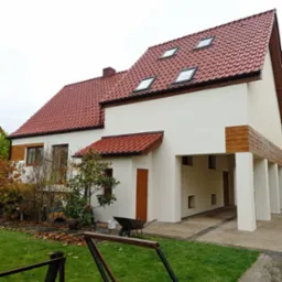 Dom jednorodzinny z czerwoną dachówką po renowacji elewacji, z widocznym podjazdem i fragmentem ogrodu. Uwagę zwraca jasna, odświeżona kolorystyka ścian.