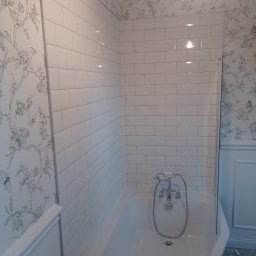 USŁUGI REMONTOWO-BUDOWLANE BIELAWSKI KRZYSZTOF MIROSŁAW - Wykończona łazienka z białą wanną, baterią i białymi, prostokątnymi płytkami na ścianach. Tapeta w kwiaty i motyle dodaje elegancji. Widoczny fragment parawanu nawannowego.