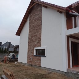 USŁUGI REMONTOWO-BUDOWLANE BIELAWSKI KRZYSZTOF MIROSŁAW - Wykończona elewacja domu jednorodzinnego z fragmentem ściany wyłożonym kamieniem dekoracyjnym i pomalowaną resztą na biało. Widoczna mała betoniarka na posesji.