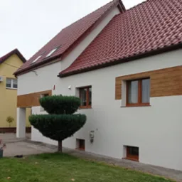 Biała elewacja domu z poziomymi pasami imitującymi drewno, brązowe okna i dach z czerwonej dachówki. Na pierwszym planie zielony krzew.