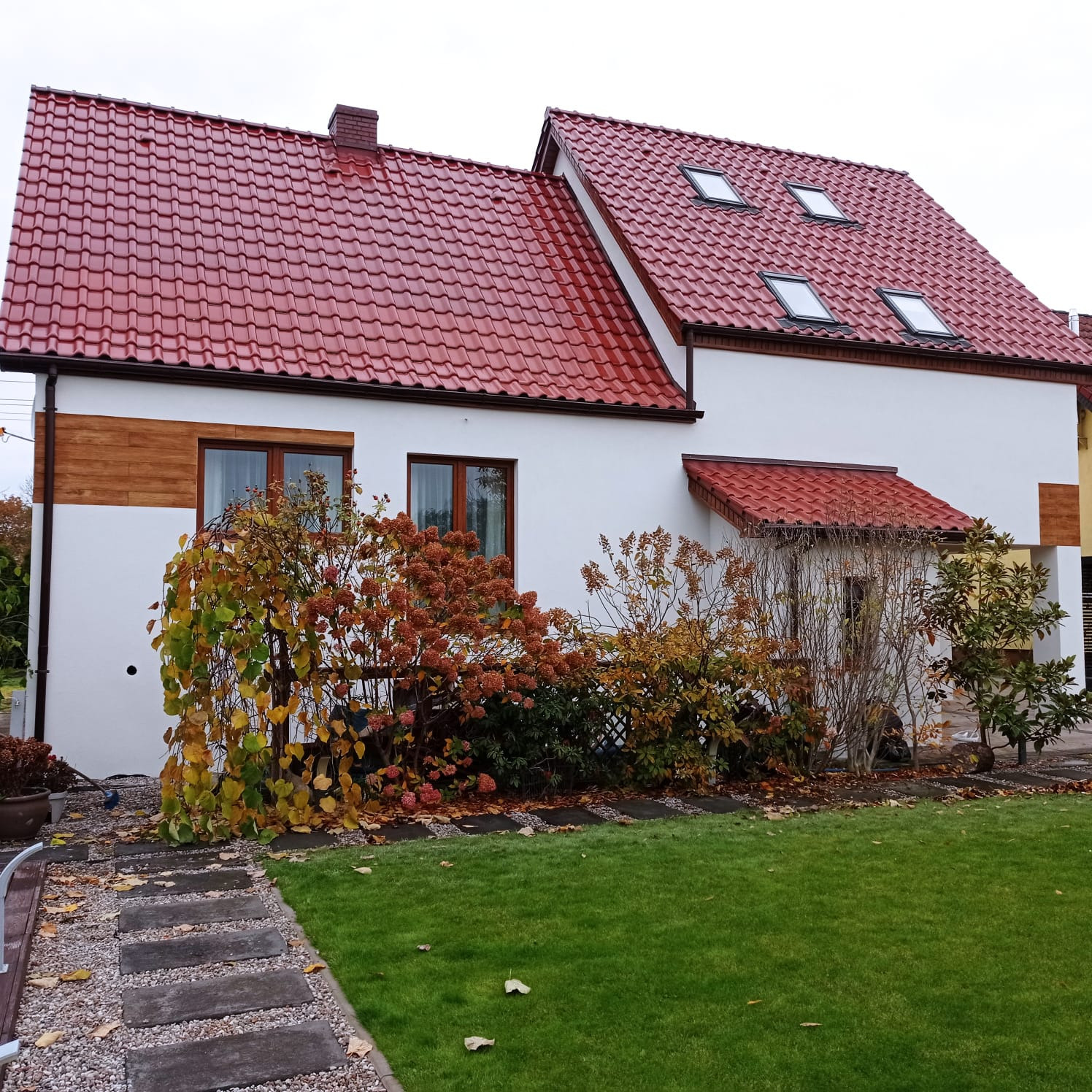 Biały dom z czerwoną dachówką i brązowymi oknami, otoczony zielonym trawnikiem i jesienną roślinnością. Fragment drewnianej elewacji po lewej stronie.