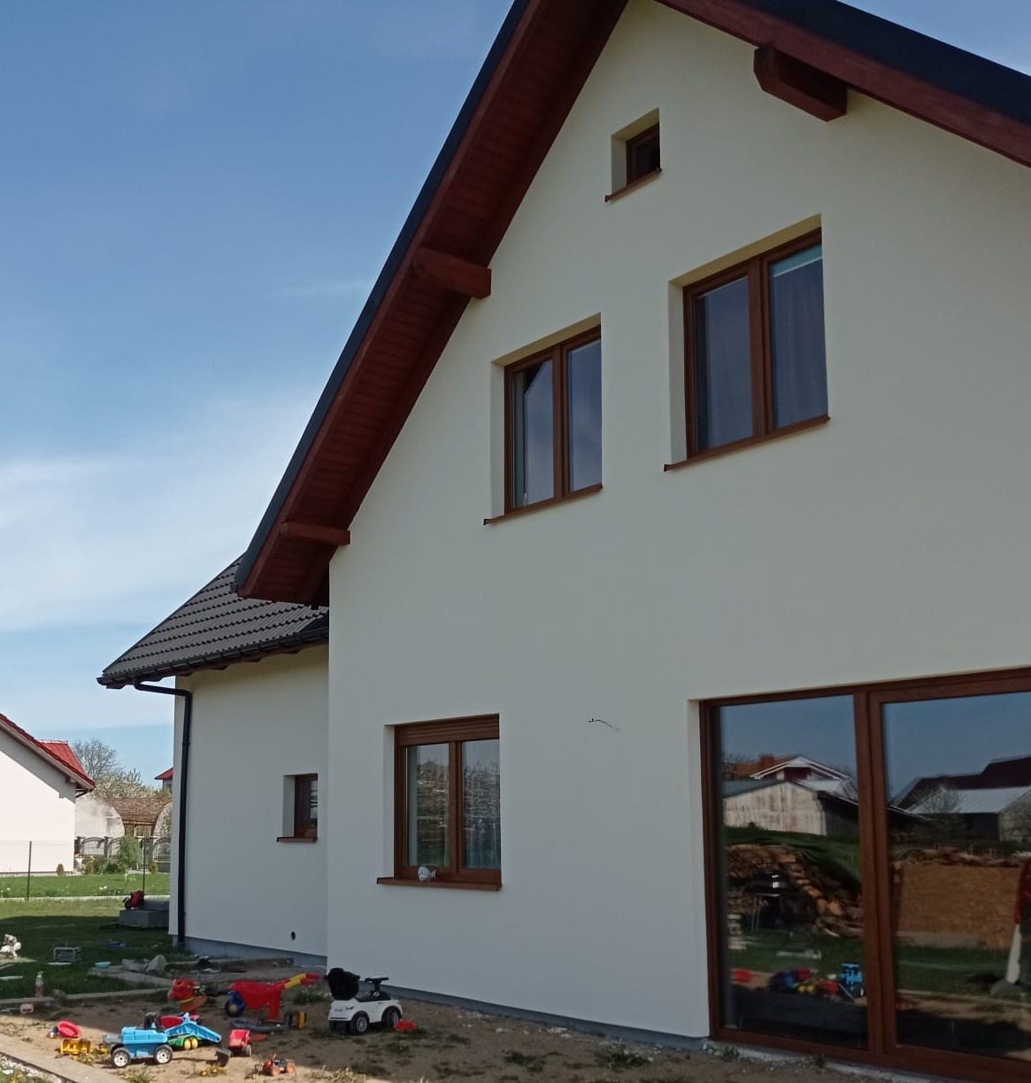 Jasna elewacja domu jednorodzinnego z brązowymi oknami i dachem, zabawki dziecięce na pierwszym planie, błękitne niebo w tle. Widok z boku, w słoneczny dzień.