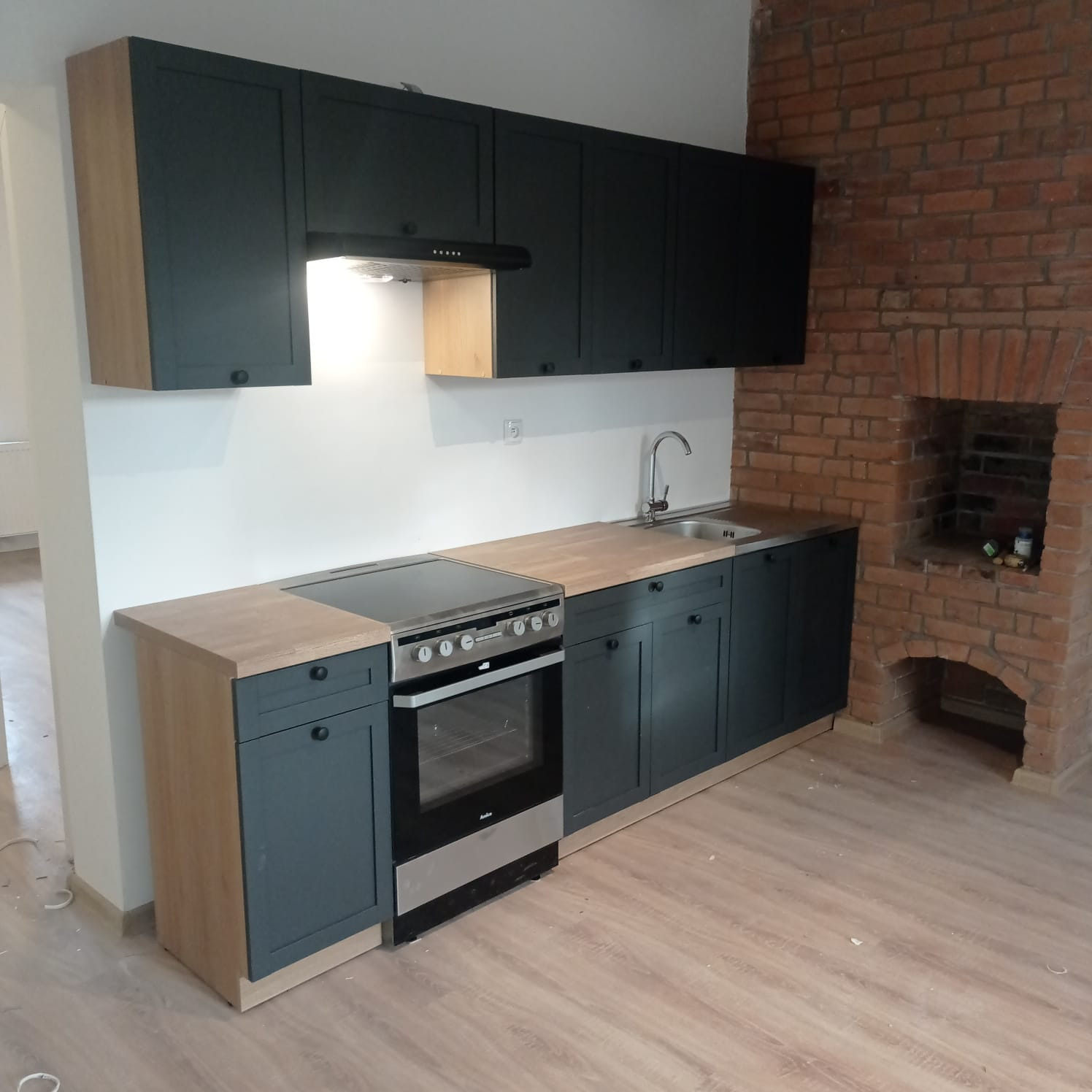 Nowoczesna kuchnia z ciemnymi szafkami, drewnianym blatem i ceglanym kominkiem w tle. Widoczna płyta indukcyjna i piekarnik.  Podłoga wyłożona panelami.