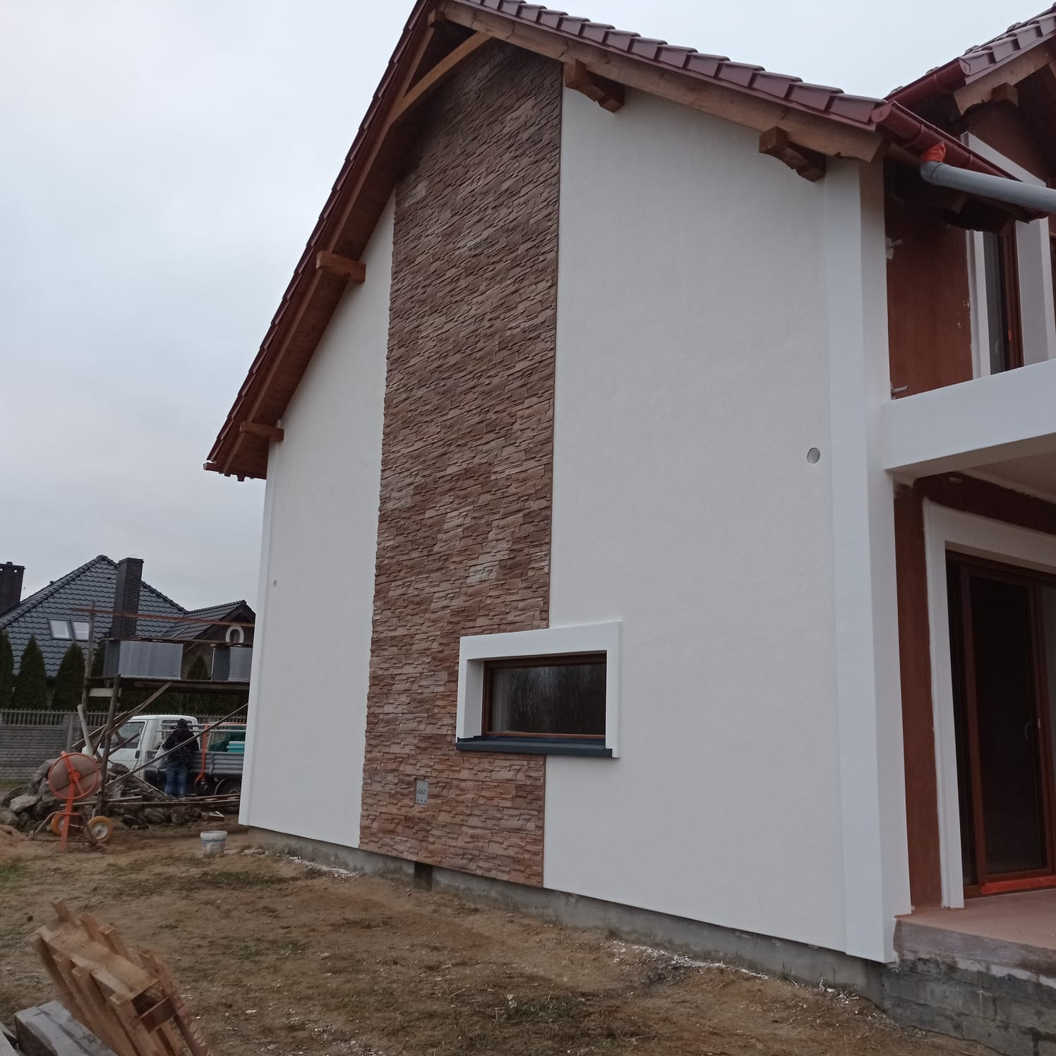 Wykończona elewacja domu jednorodzinnego z fragmentem ściany wyłożonym kamieniem dekoracyjnym i pomalowaną resztą na biało. Widoczna mała betoniarka na posesji.