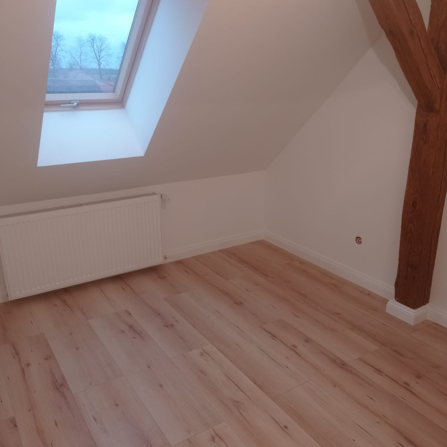 Jasne poddasze z oknem dachowym, drewnianą belką i panelami podłogowymi. Świeżo pomalowane ściany w odcieniach bieli, nowy grzejnik. Widok na zewnątrz przez okno.