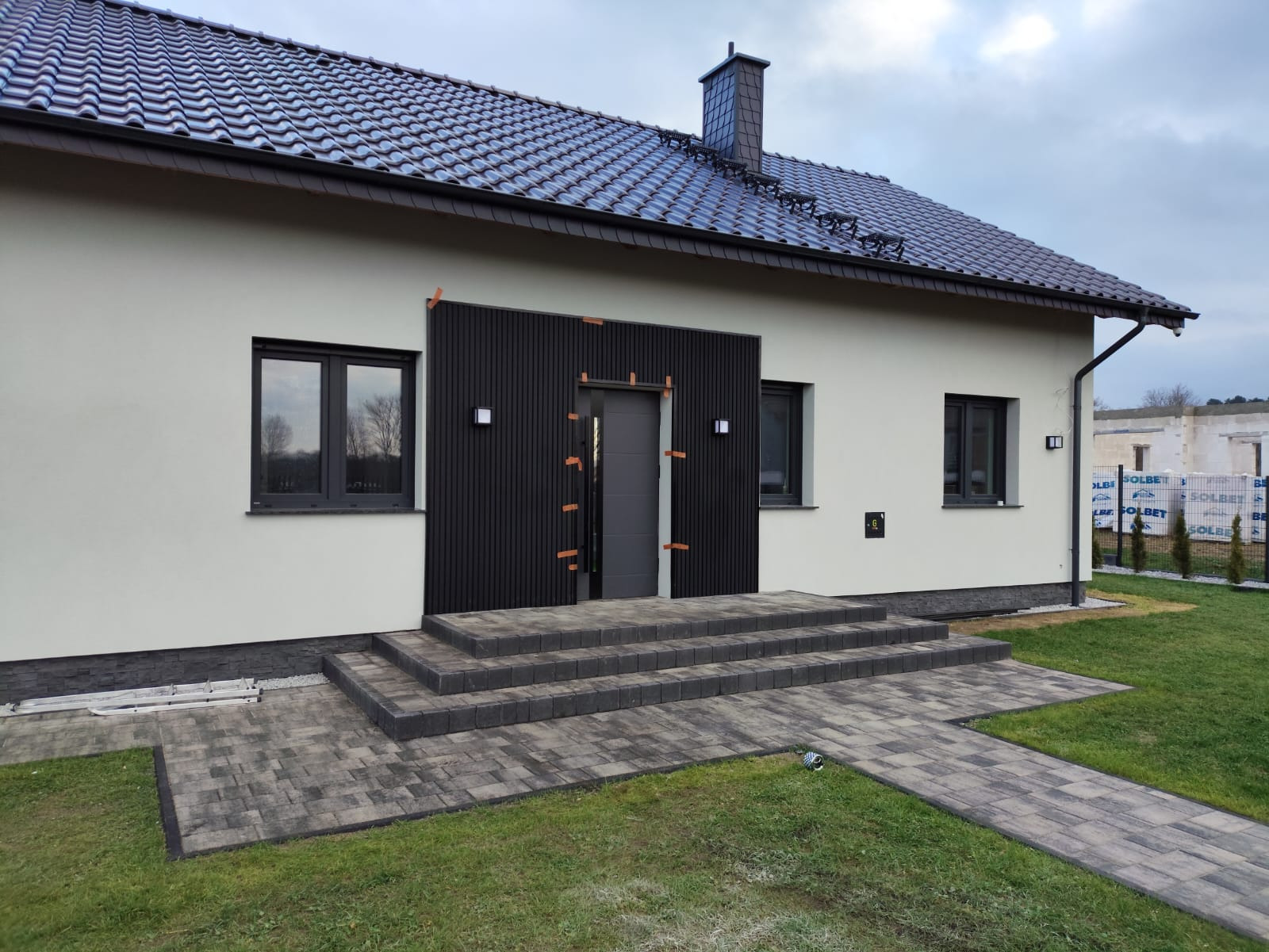 Nowoczesny dom z jasną elewacją, ciemnym panelem przy wejściu i schodami z kostki. Widoczny fragment trawnika i ogrodzenia. Detal: czarny panel częściowo zabezpieczony taśmą.