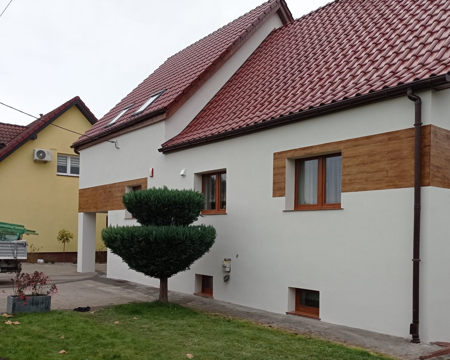 Biała elewacja domu z poziomymi pasami imitującymi drewno, brązowe okna i dach z czerwonej dachówki. Na pierwszym planie zielony krzew.