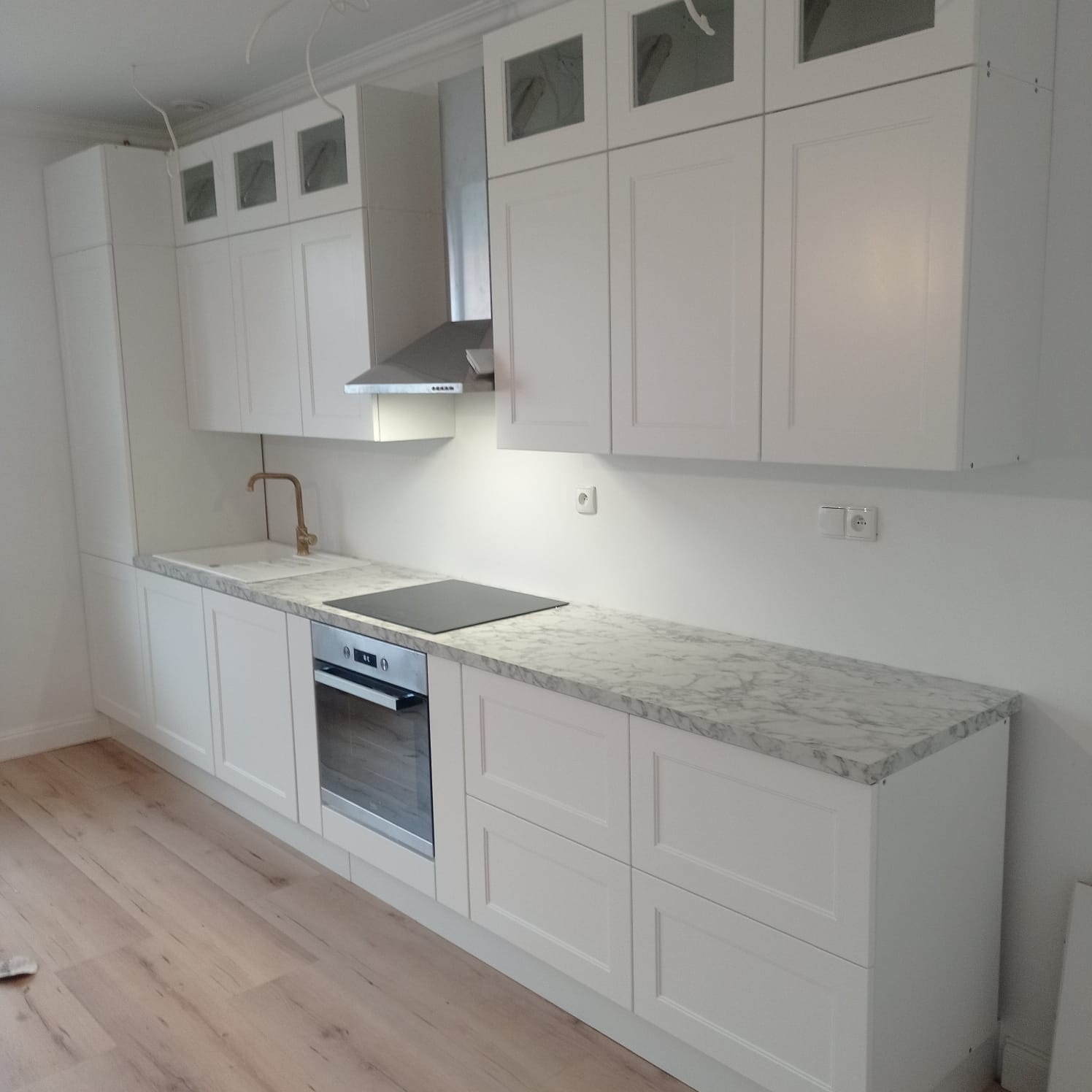 Biała, nowoczesna kuchnia z marmurowym blatem i złotą armaturą. Górne szafki częściowo przeszklone. Widoczne podłączenia elektryczne do oświetlenia.