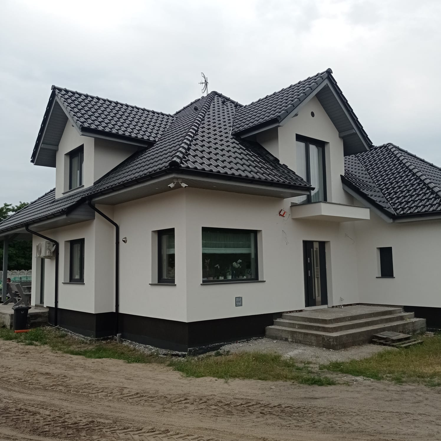 Dwukondygnacyjny dom z białą elewacją i czarnym dachem z dachówki. Widoczne czarne okna i drzwi. Nieukończone schody wejściowe. Na dachu widoczna antena.