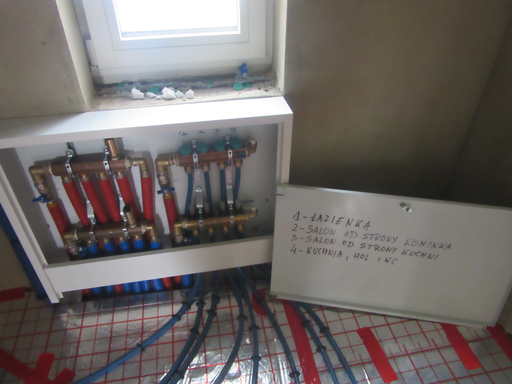 Rozdzielacz ogrzewania podłogowego w białej obudowie, z widocznymi rurami w kolorach czerwonym i niebieskim, ułożonymi na folii izolacyjnej. Obok tablica z opisem.