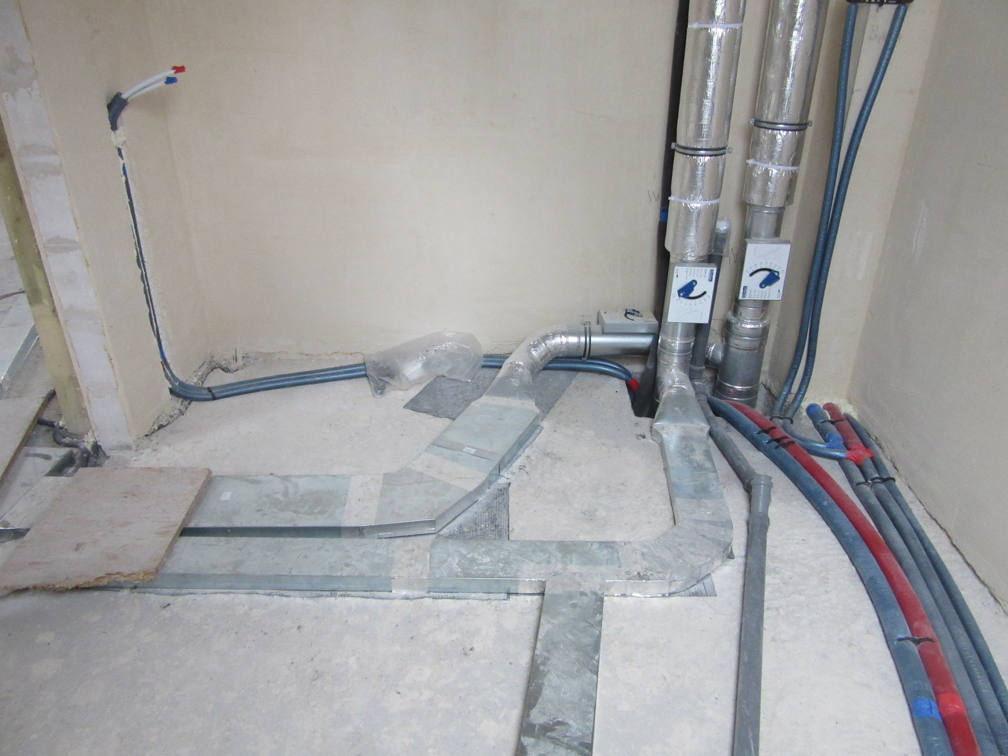 Instalacja grzewcza w trakcie montażu: rury w izolacji, kanały wentylacyjne i przewody ułożone na podłodze, widoczne zawory. Etap prac wykończeniowych.