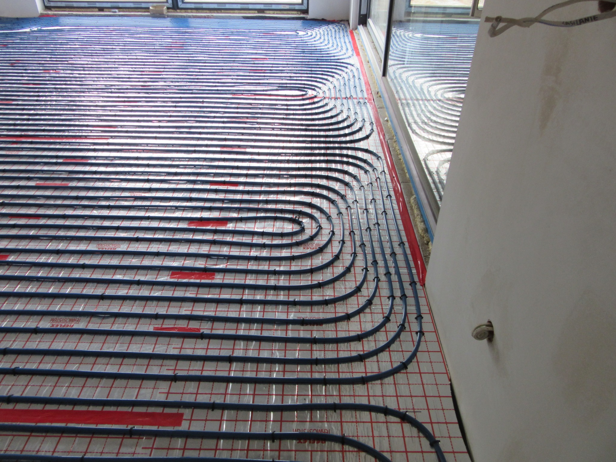 Ułożone rury ogrzewania podłogowego na folii refleksyjnej z czerwoną siatką, widok z góry, przy ścianie wystający zawór.