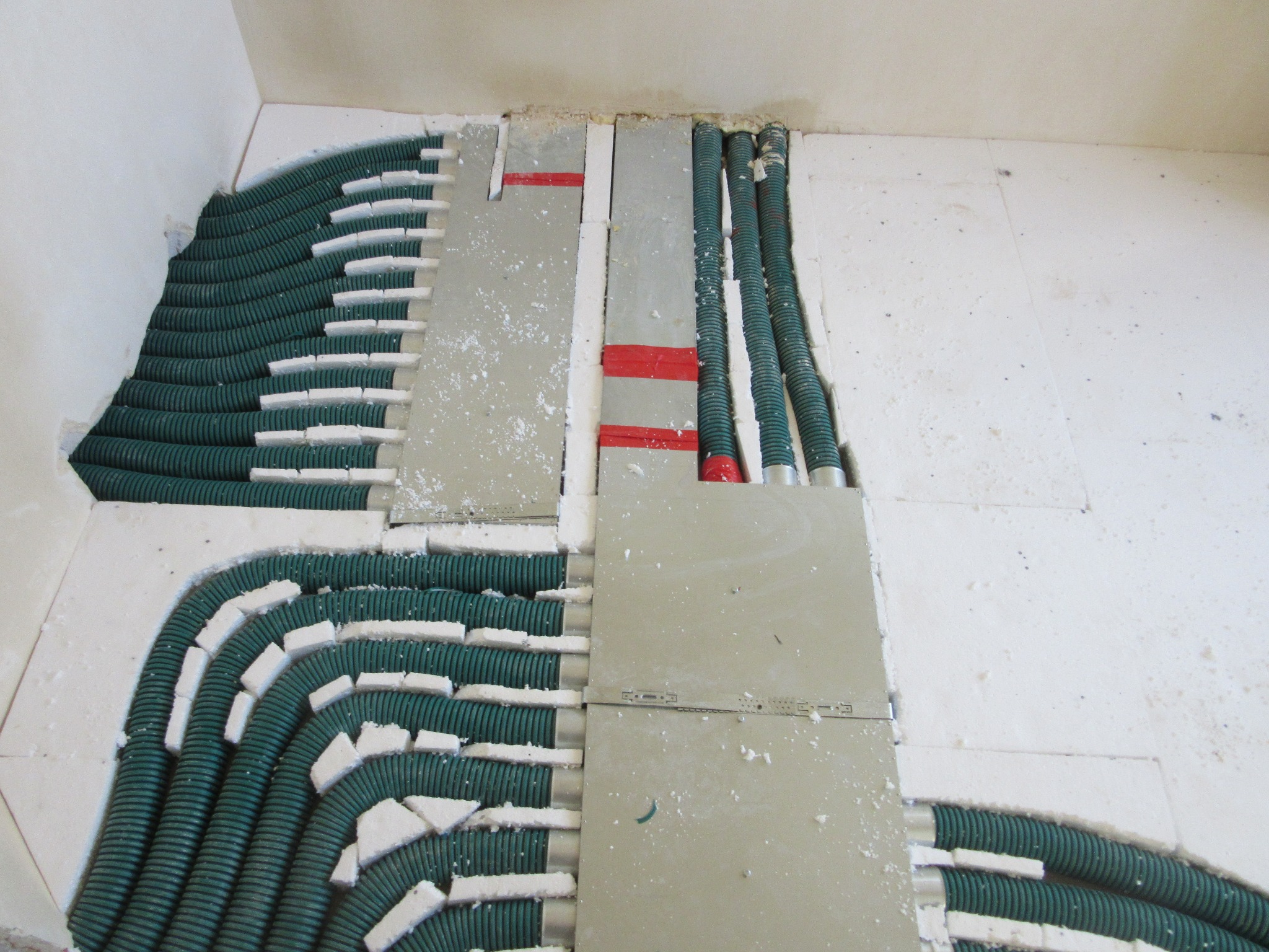 Zielone rury ogrzewania podłogowego w styropianowej izolacji, częściowo zakryte szarą płytą. Widoczne czerwone taśmy. Ujęcie z góry, w trakcie instalacji.