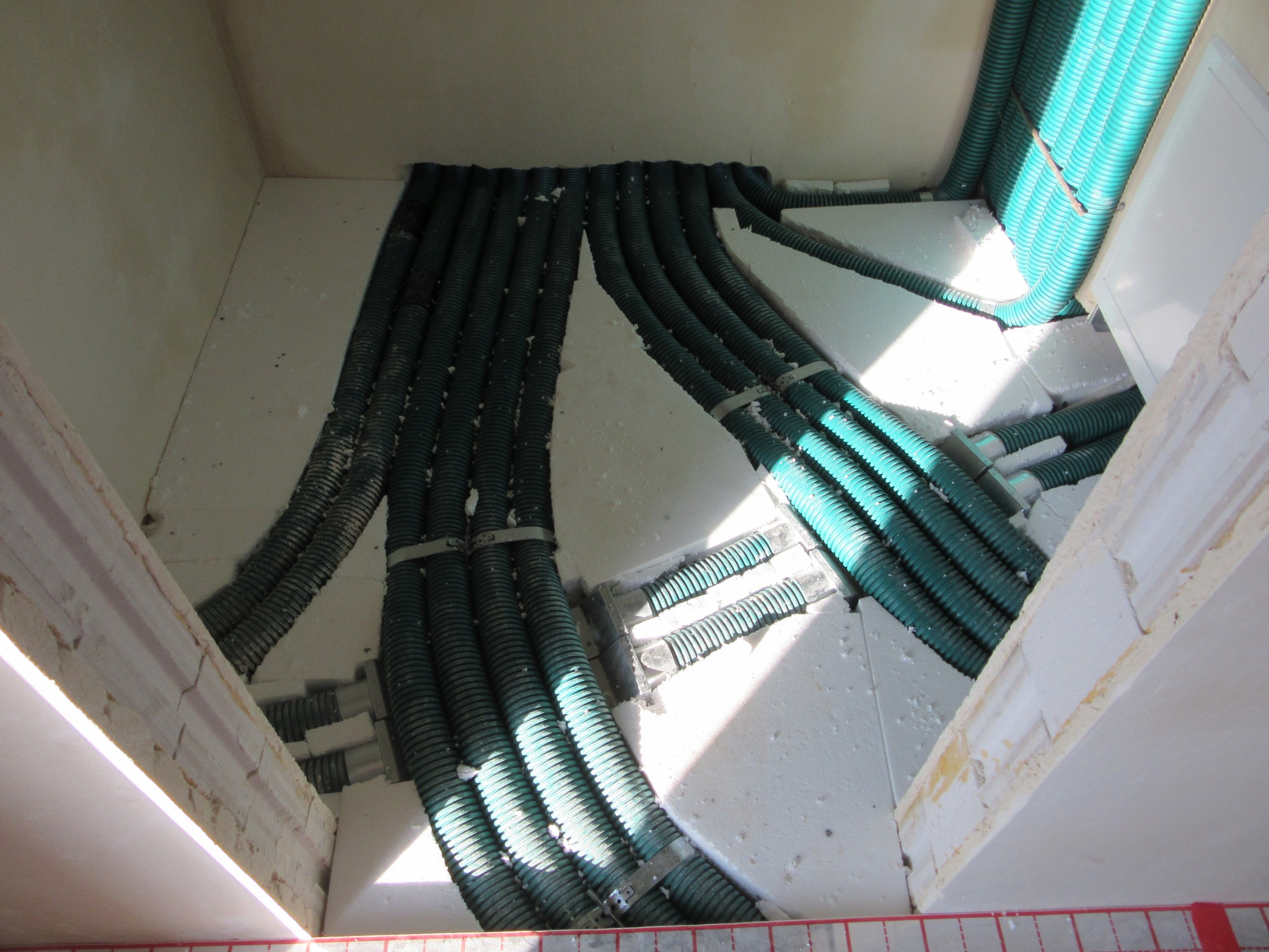 Zielone rury instalacji ogrzewania podłogowego ułożone w białym styropianie, zabezpieczone metalowymi klamrami, widok z góry w surowym wnętrzu budynku.