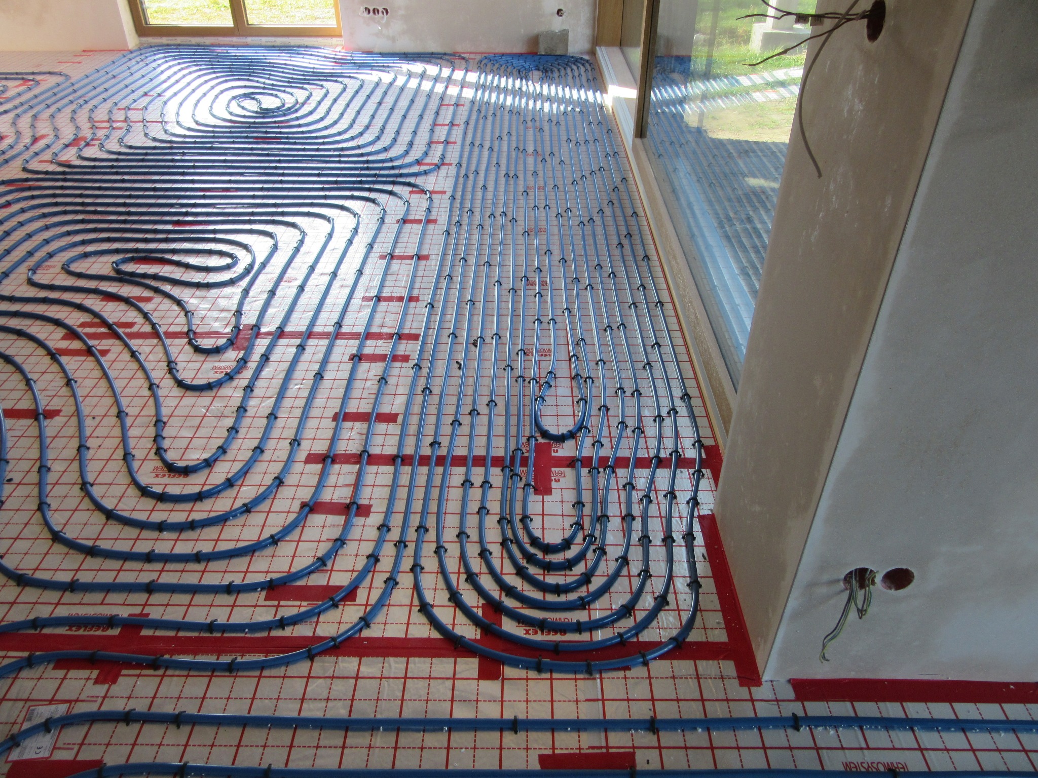 Niebieskie rury ogrzewania podłogowego ułożone na czerwonej folii z nadrukiem, widoczne okno i fragment ściany z wystającymi kablami.