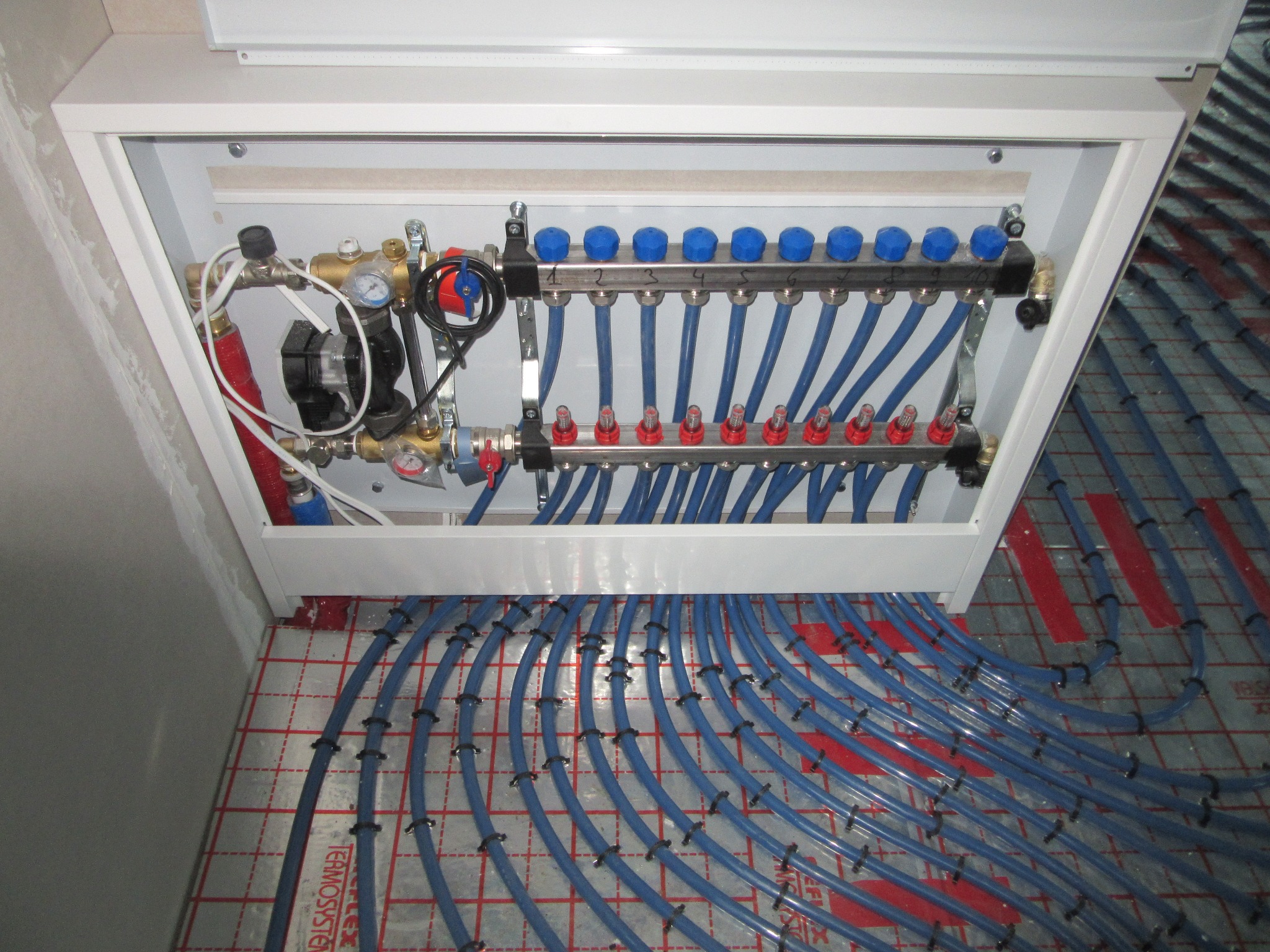 Rozdzielacz ogrzewania podłogowego z niebieskimi rurami na macie izolacyjnej z czerwonym nadrukiem, umieszczony w białej szafce wnękowej.