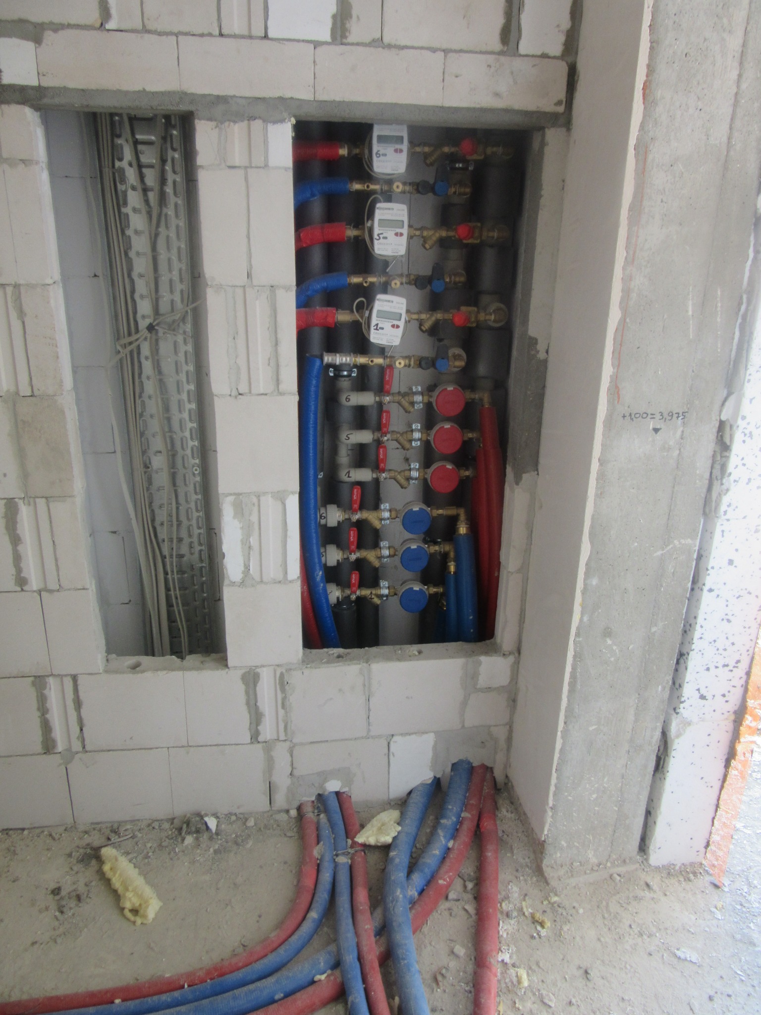 Rozdzielacz ogrzewania podłogowego z licznikami ciepła, zamontowany w ścianie z betonu komórkowego, w trakcie prac instalacyjnych w Warszawie. Rury czerwone i niebieskie.