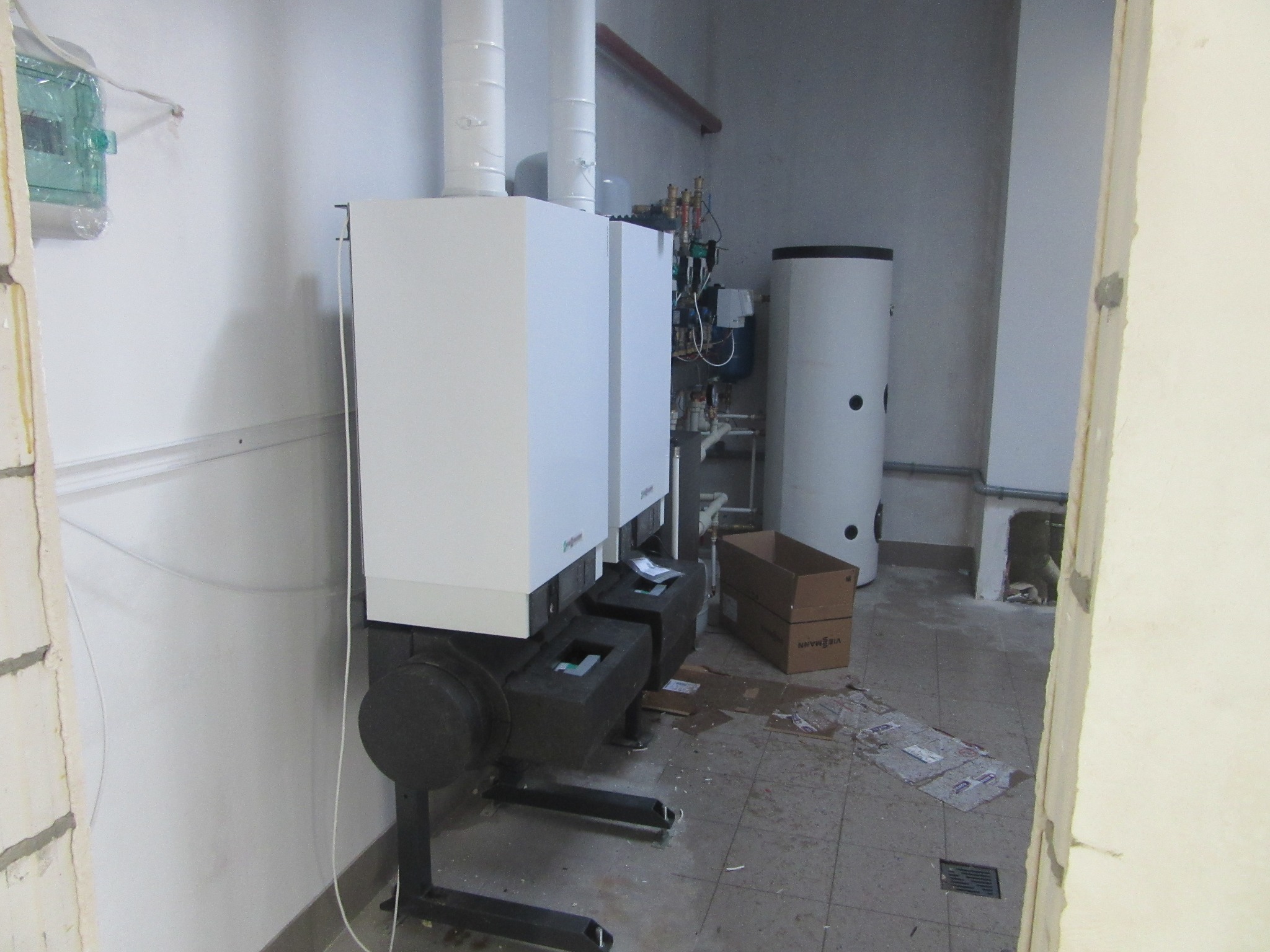 Nowoczesny, biały piec gazowy Viessmann z czarnymi elementami, zainstalowany w surowym pomieszczeniu z widocznymi instalacjami i bojlerem. Widok zza framugi.