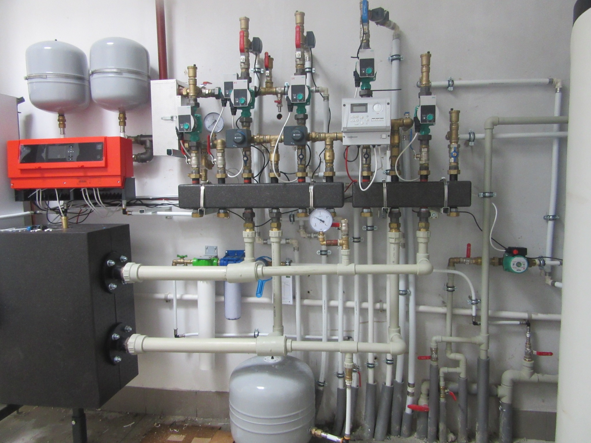 Złożona instalacja grzewcza z licznymi rurami, zaworami, manometrami i elementami sterującymi, zamontowana na ścianie. Widoczny piec i zbiorniki wyrównawcze.