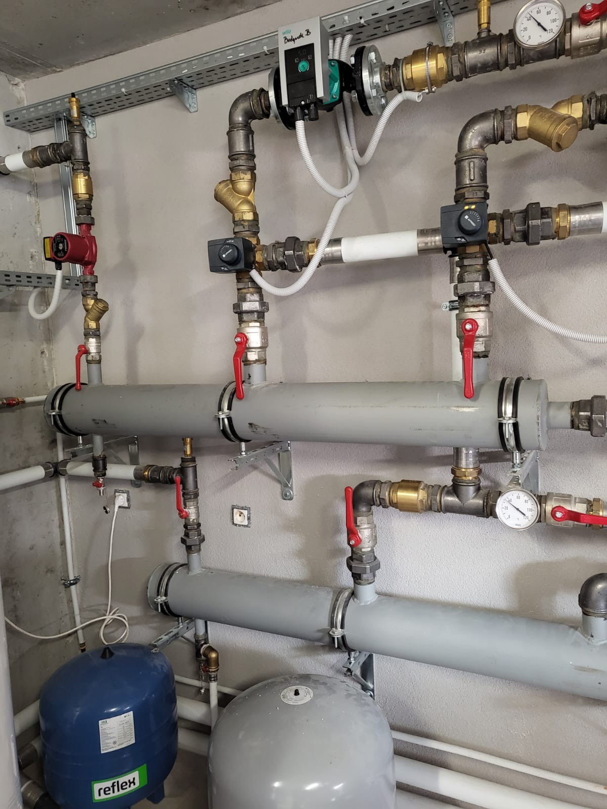 Szczegółowe ujęcie instalacji grzewczej z szarymi rurami, zaworami z czerwonymi uchwytami, manometrami i pompą obiegową, zamontowanej na szarej ścianie.