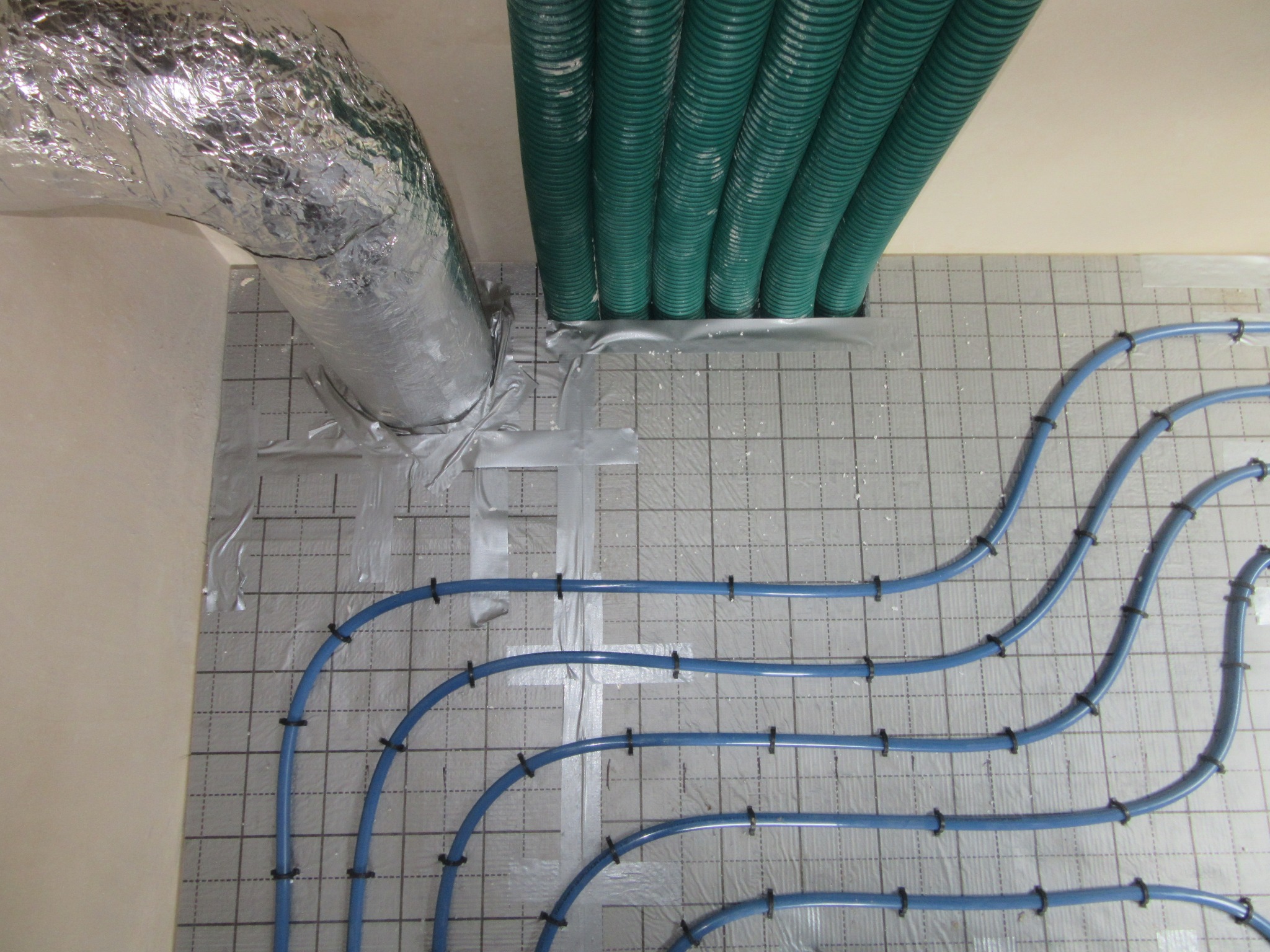 Niebieskie rury ogrzewania podłogowego, zamocowane na izolacji z widoczną siatką, z zielonymi rurami wentylacyjnymi i srebrnym kanałem wentylacyjnym.