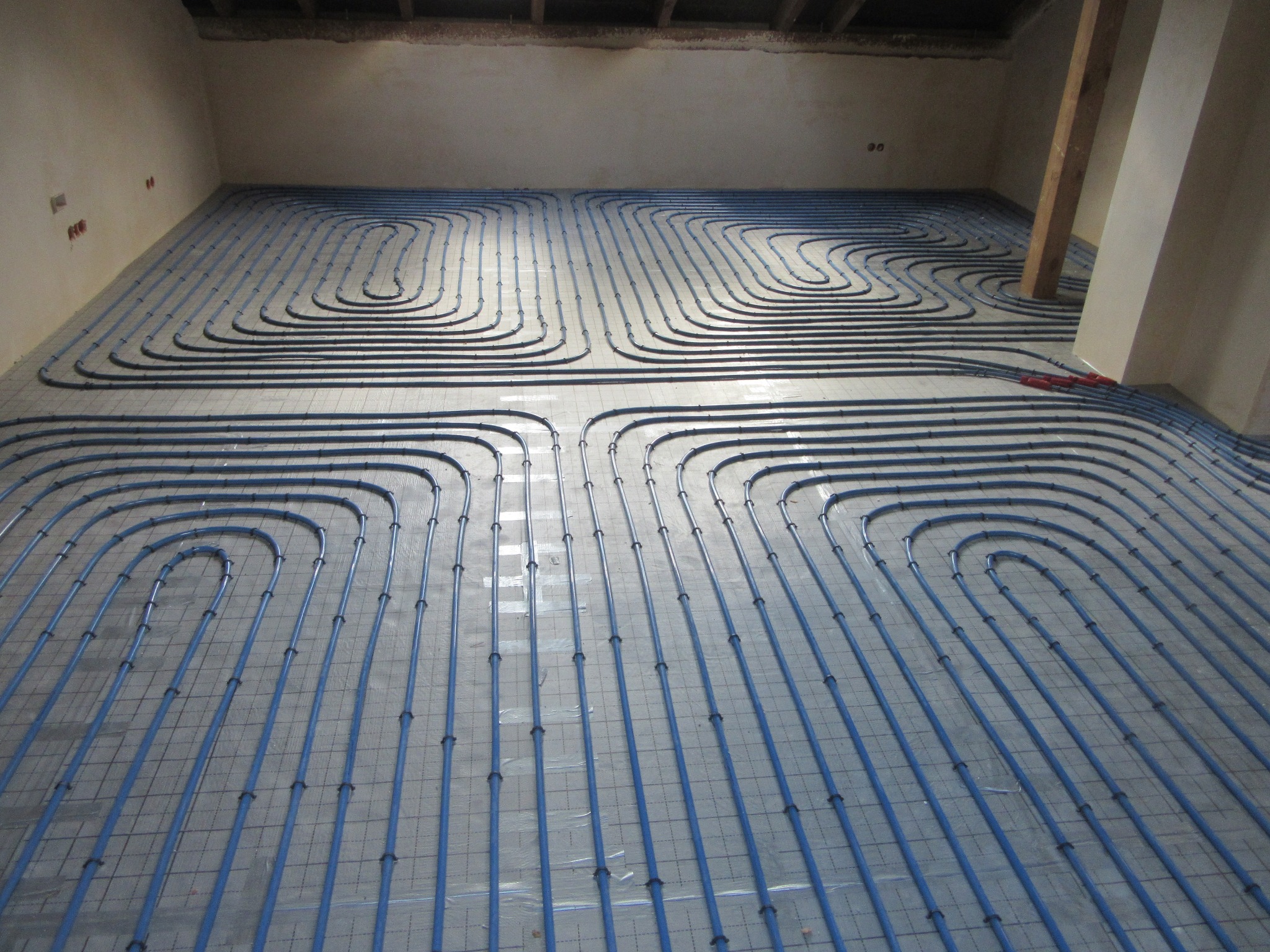 Niebieskie rury ogrzewania podłogowego ułożone na szarej macie w surowym wnętrzu. Widoczne mocowania rur i fragment ściany z instalacją elektryczną.